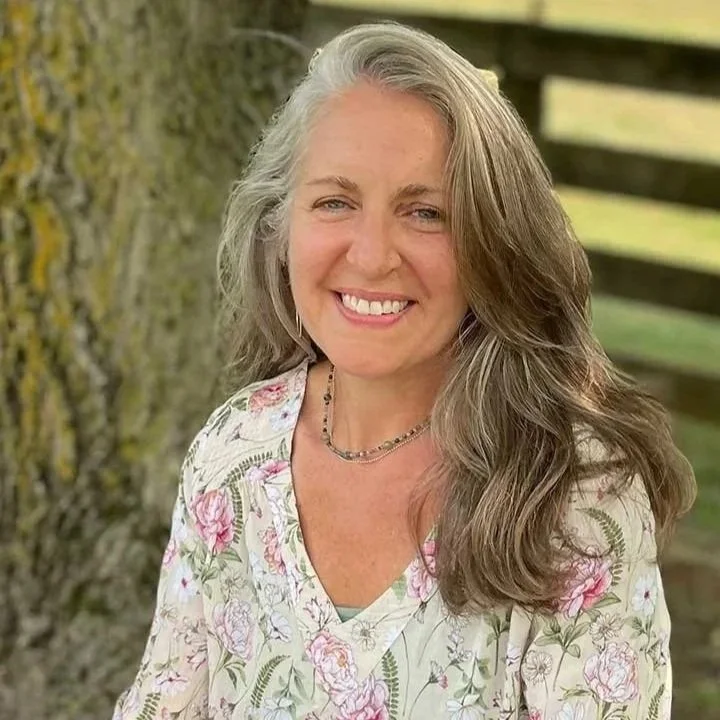 A woman with long, wavy gray hair smiling outdoors near a tree and a wooden fence, wearing a floral blouse and a necklace.