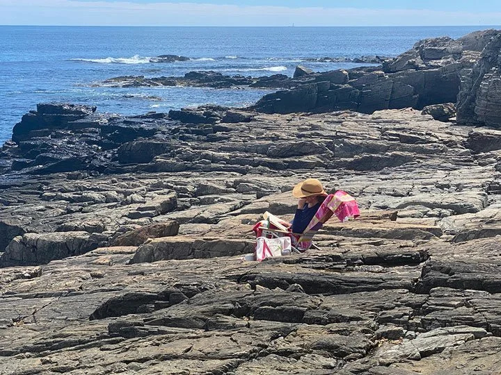 Person sitting on rocky shoreline reading a book, wearing a sun hat, with a pink towel and beach bag nearby, overlooking the ocean.