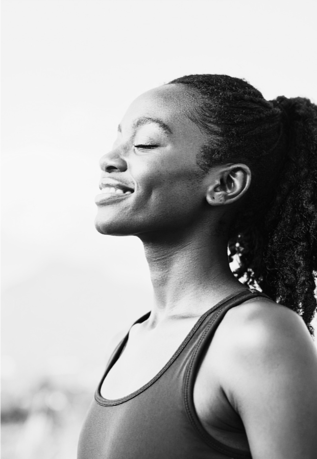 A black and white portrait of a woman with her eyes closed and a peaceful smile, wearing a sports top and outdoors.