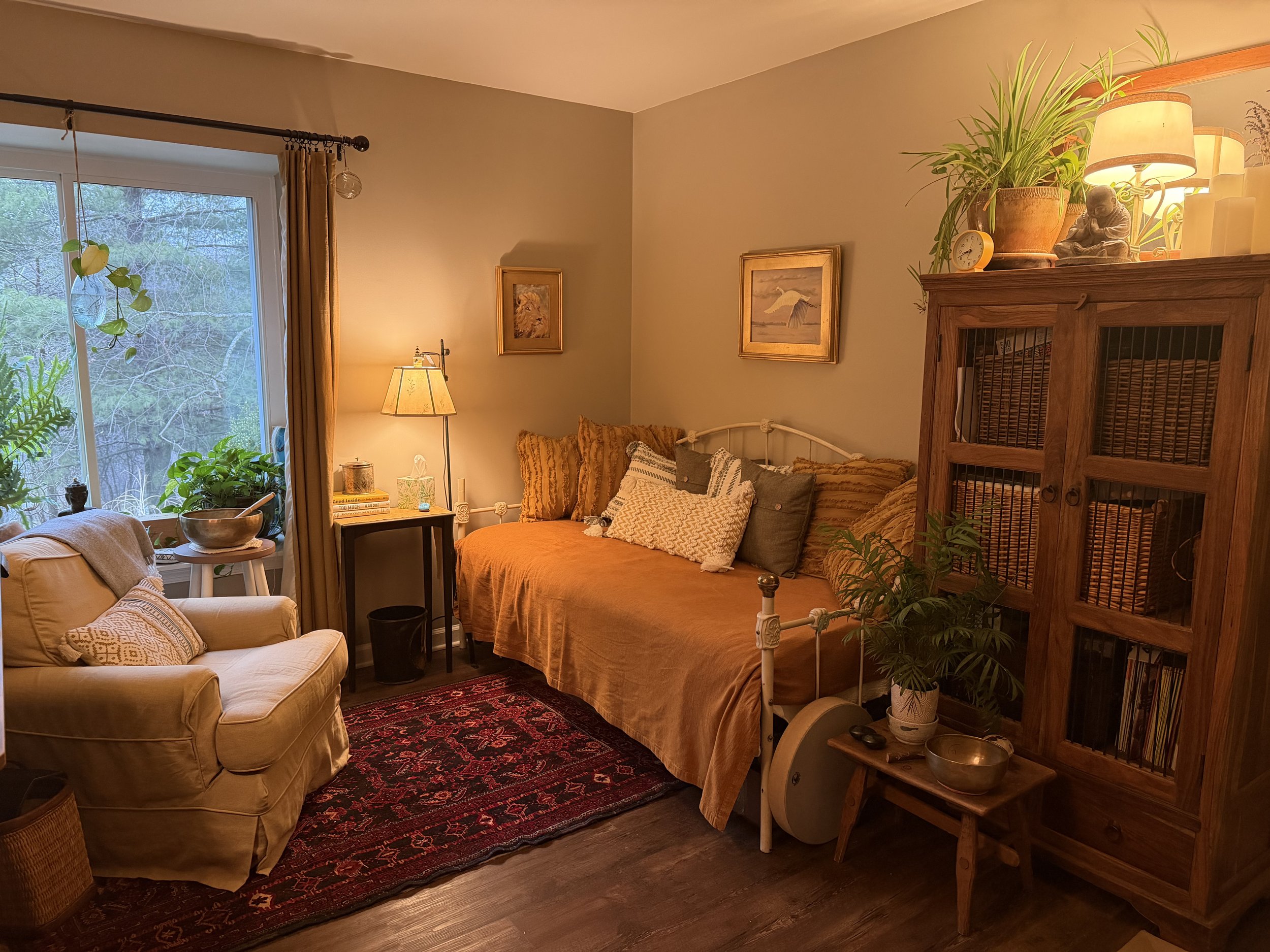 A cozy living room corner with a yellow armchair, a beige couch, a small black side table with a lamp, plants, framed artwork, a wooden cabinet, and decorative items, illuminated by warm lighting.