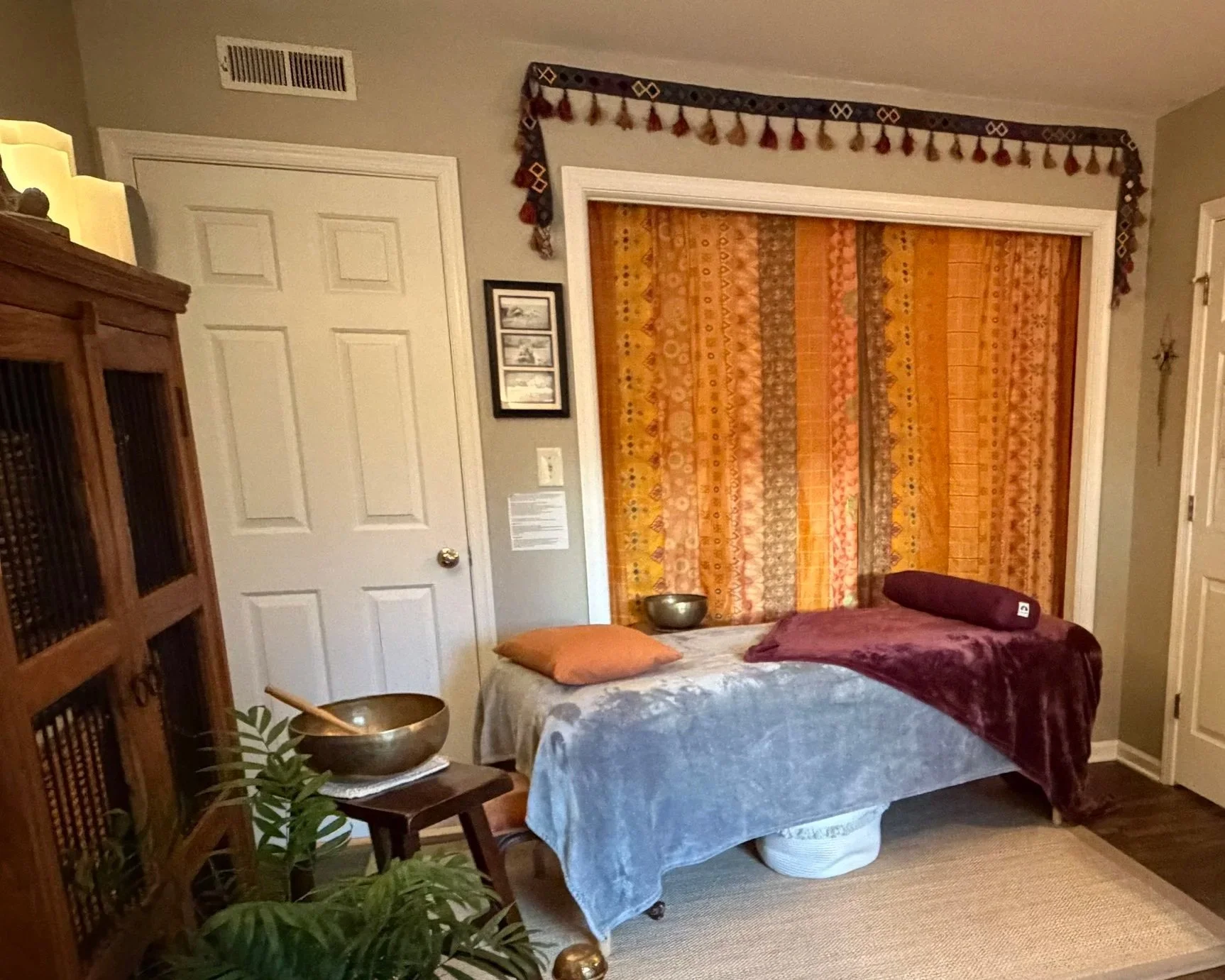 A massage room with a massage table covered in gray and maroon blankets, decorative curtains in warm tones behind, and various decor including bowls, pillows, and a wooden cabinet. There's a plant in the foreground.