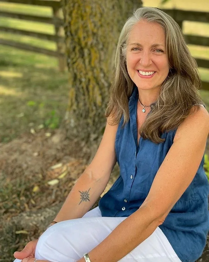 A woman with long gray hair, wearing a sleeveless blue shirt and white pants, sitting outdoors in front of a tree with a wooden fence in the background. She is smiling and looking at the camera, with a tattoo of a sun on her left arm.