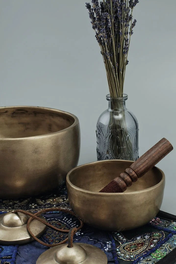 Two brass singing bowls, a glass vase with lavender flowers, a small bell with a leather cord, and a wooden mallet on a richly embroidered blue cloth.