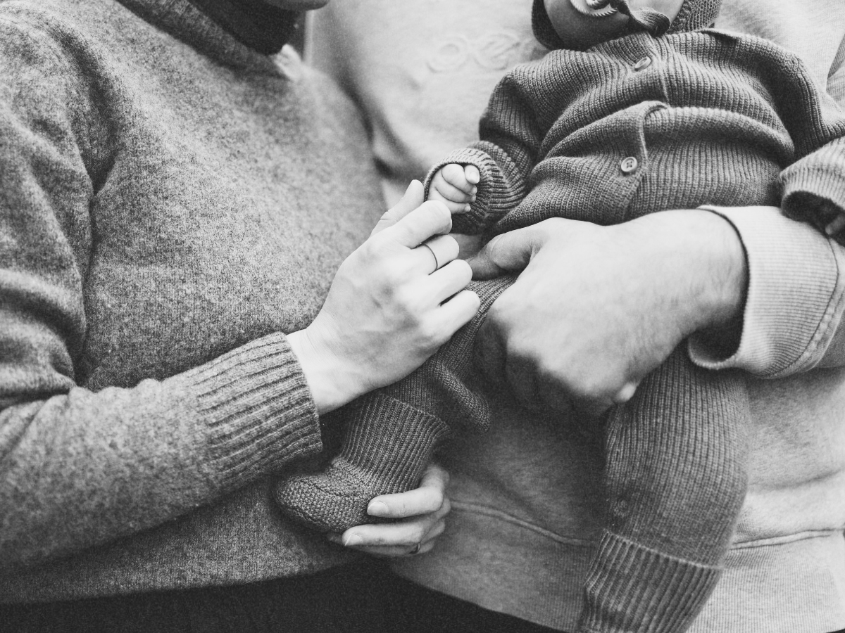 A person holding a baby's hand, with the baby sitting on someone's lap, both wearing sweaters.