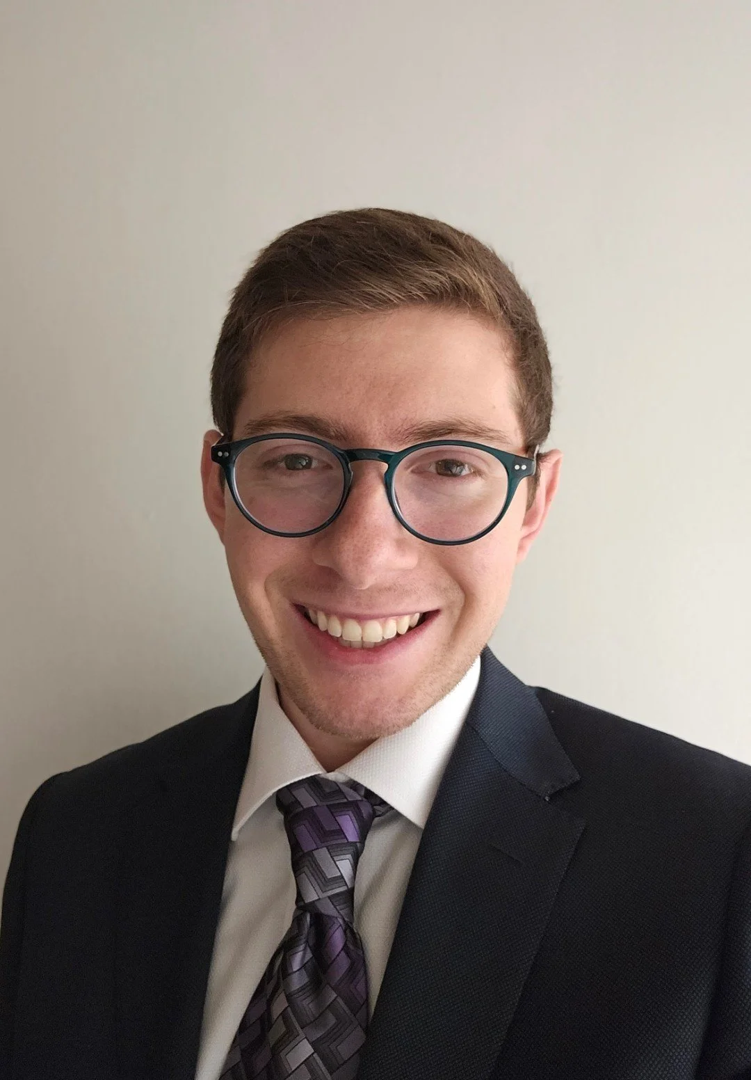 A young man with brown hair, wearing glasses, a dark blazer, a white shirt, and a patterned tie, smiling against a plain white wall.
