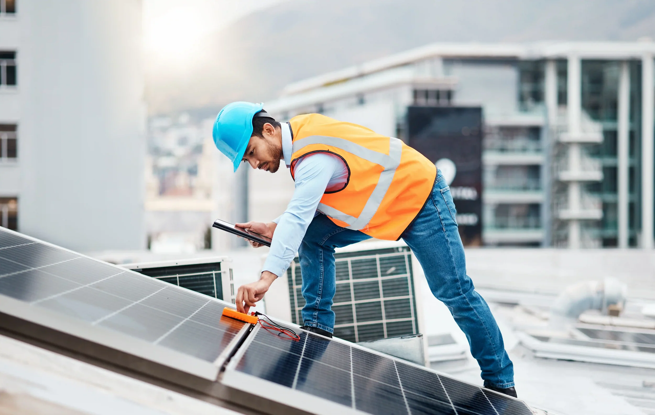 A technician wearing a blue helmet and orange safety vest inspecting solar panels on a rooftop.