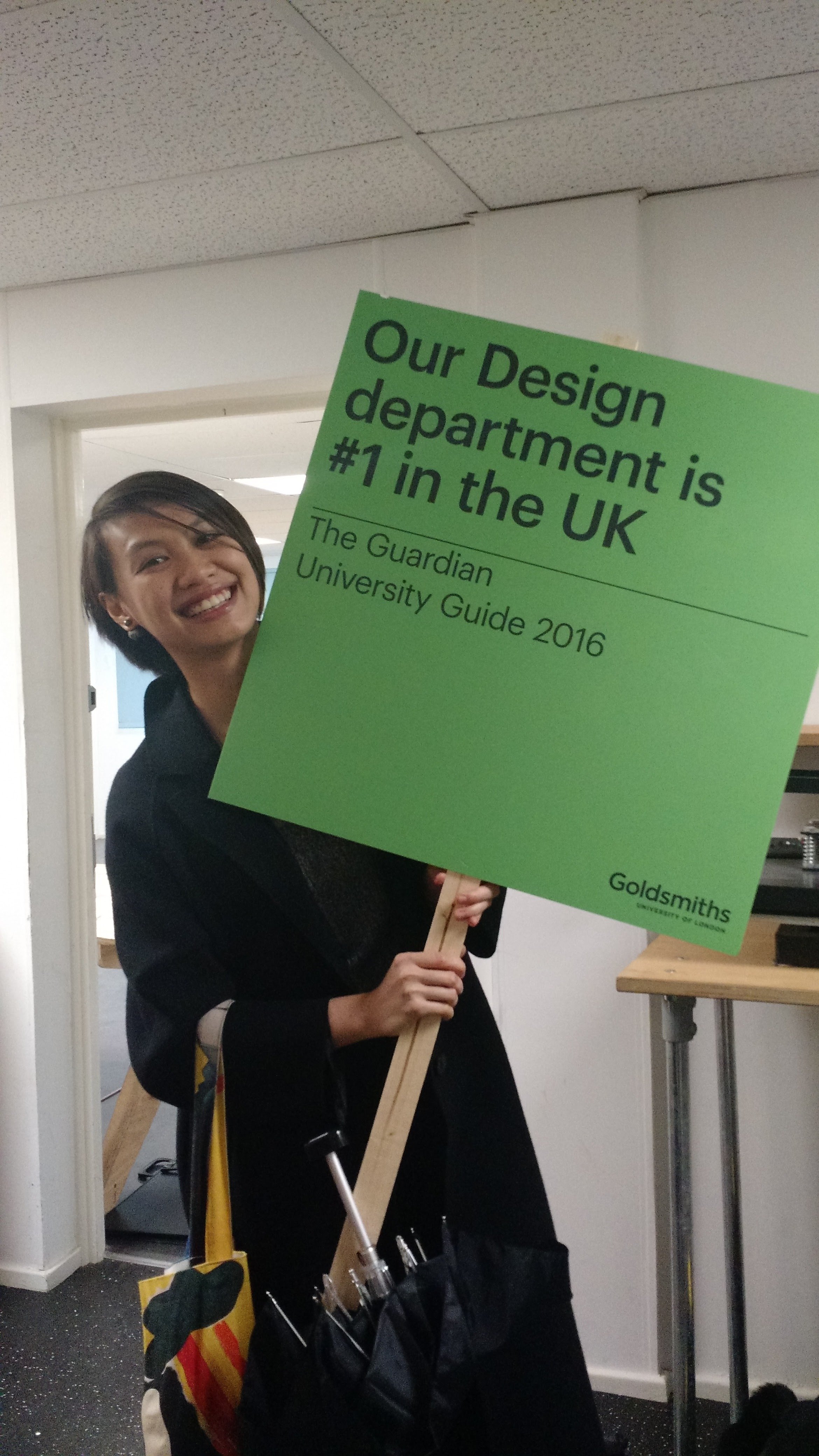 A woman smiling and holding a green sign that says 'Our Design department is #1 in the UK' and 'The Guardian University Guide 2016'.