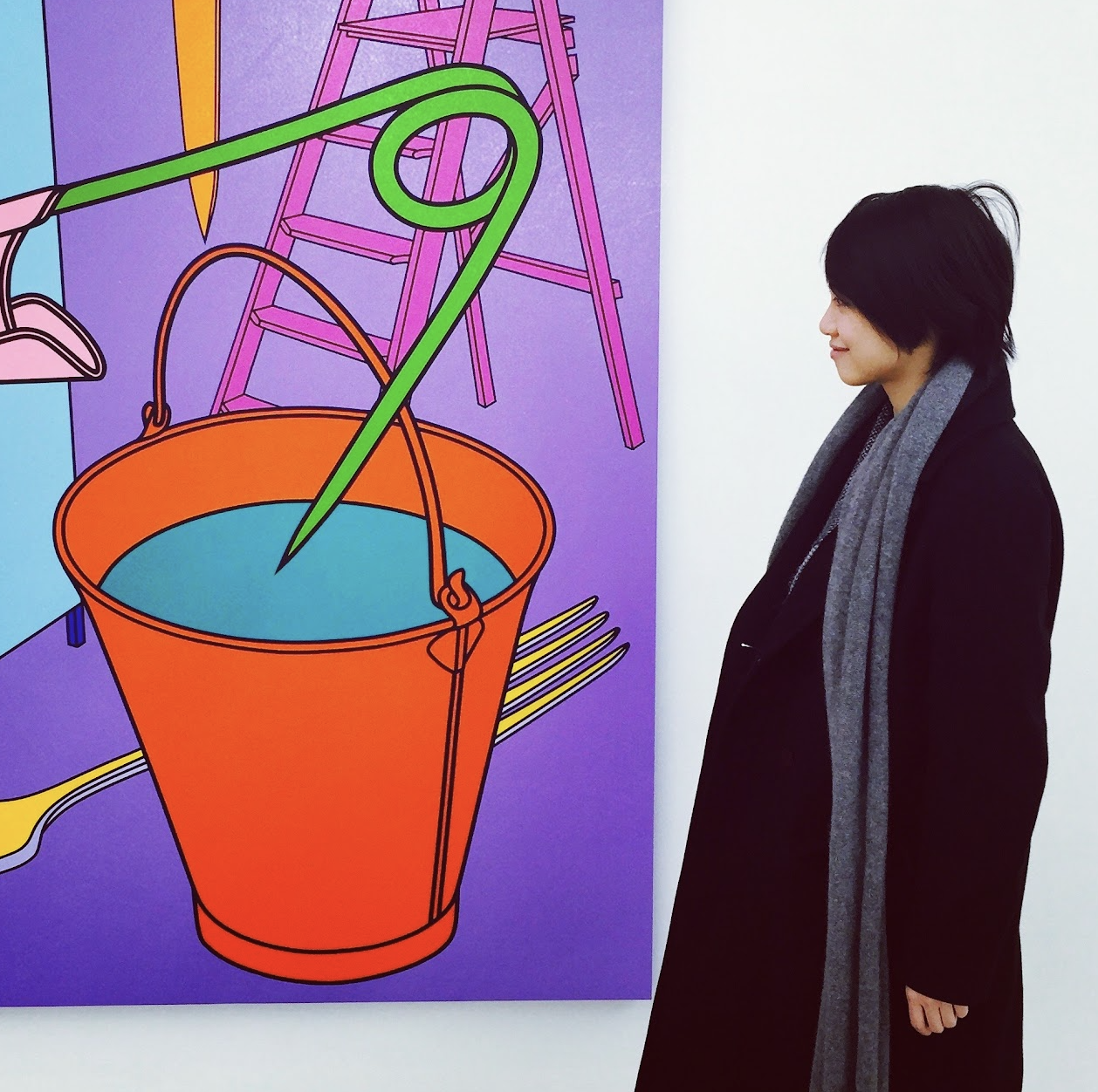 A woman with short black hair wearing a black coat and gray scarf standing and looking at a colorful abstract painting with a large orange bucket and green hook.