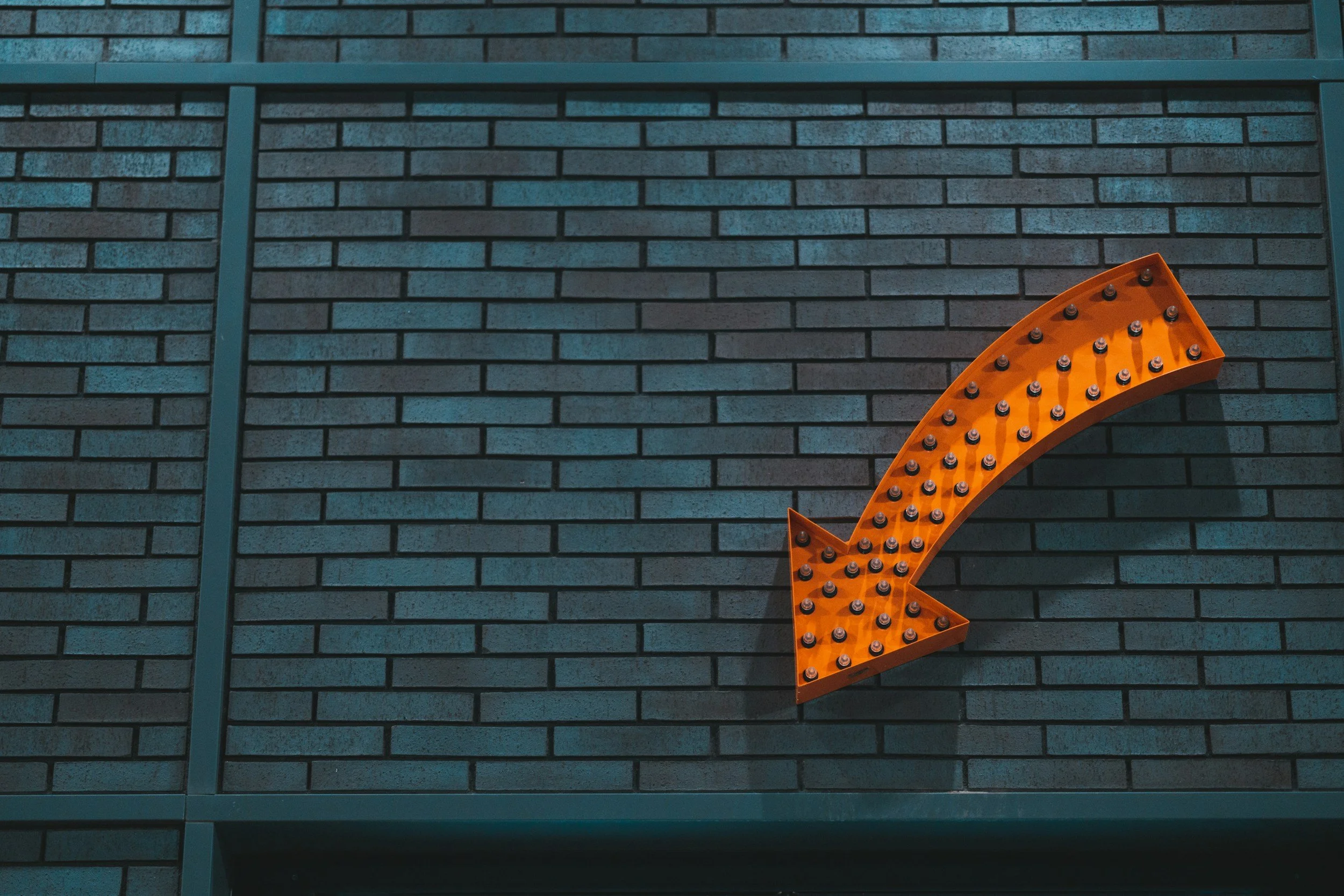 A large, orange, arrow-shaped sign with small light bulbs pointing downward on a blue brick wall.