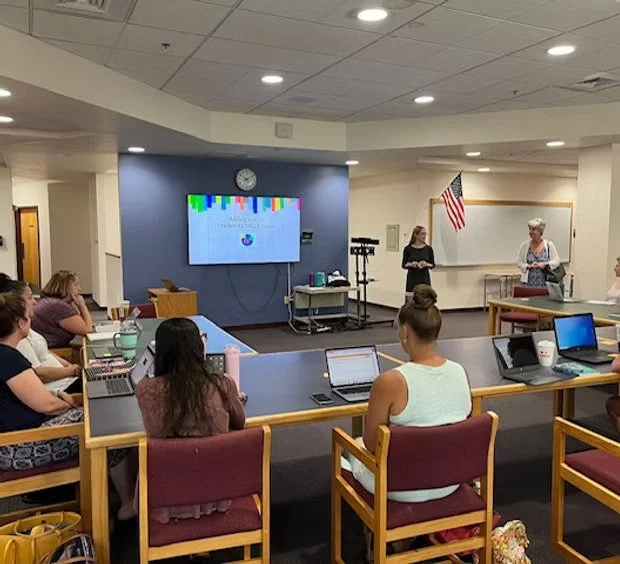 A classroom or training room with adults seated at tables using laptops, facing a speaker and a woman standing near a whiteboard, with an American flag, a large screen, and a clock on the wall.