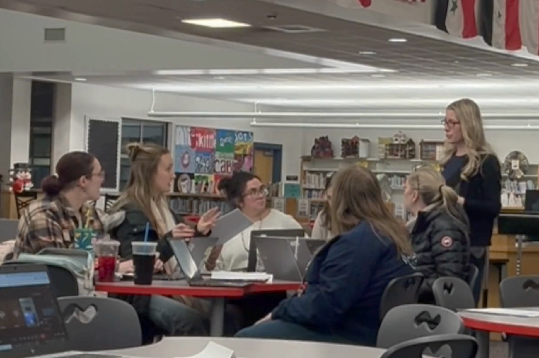 A group of women are sitting around a table in a library, listening to a woman standing and speaking. There are bookshelves, a colorful poster, and school supplies visible in the background.