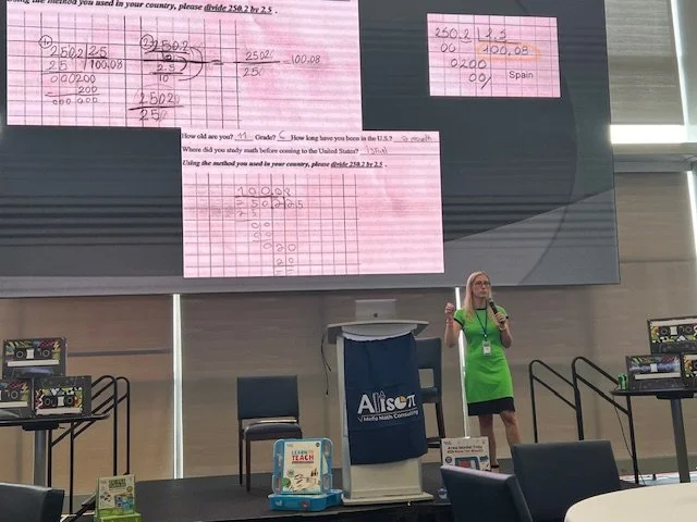 A woman in a green apron standing on a stage in front of a large screen displaying math problem solutions written on pink paper, with tables, chairs, and tables in the room.