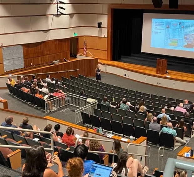 A speaker presenting to an audience in a conference room with multiple rows of seating, a large screen displaying a presentation, and an American flag in the background.