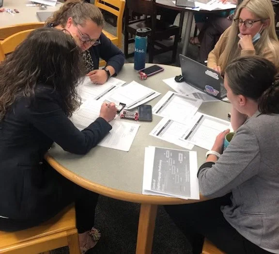 Four women sitting around a table, discussing documents and working on laptops.