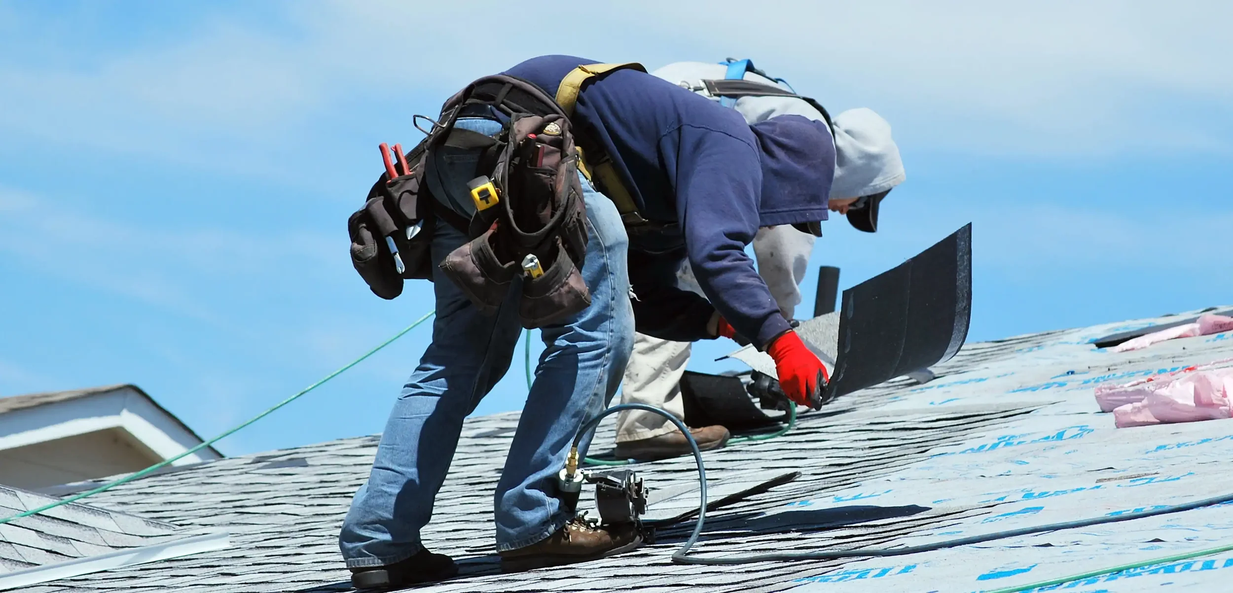 Two workers roofing a house, wearing safety gear, on a sunny day.