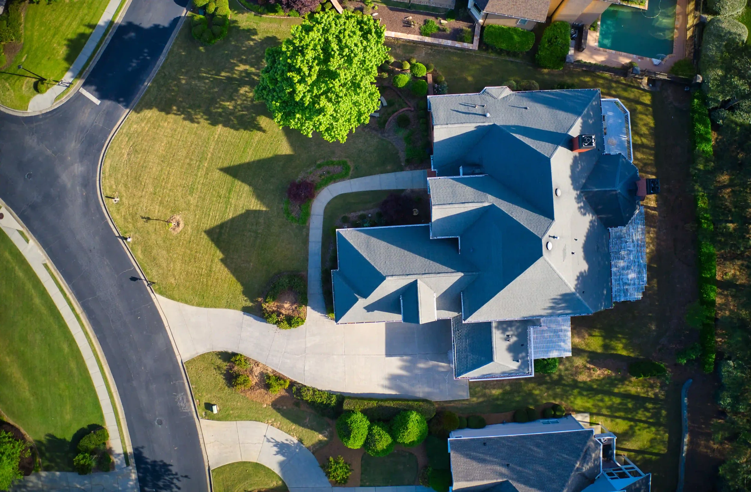 Aerial view of a house with a gray roof, surrounded by a green lawn, trees, and a driveway, with neighboring houses and swimming pools visible in the background.