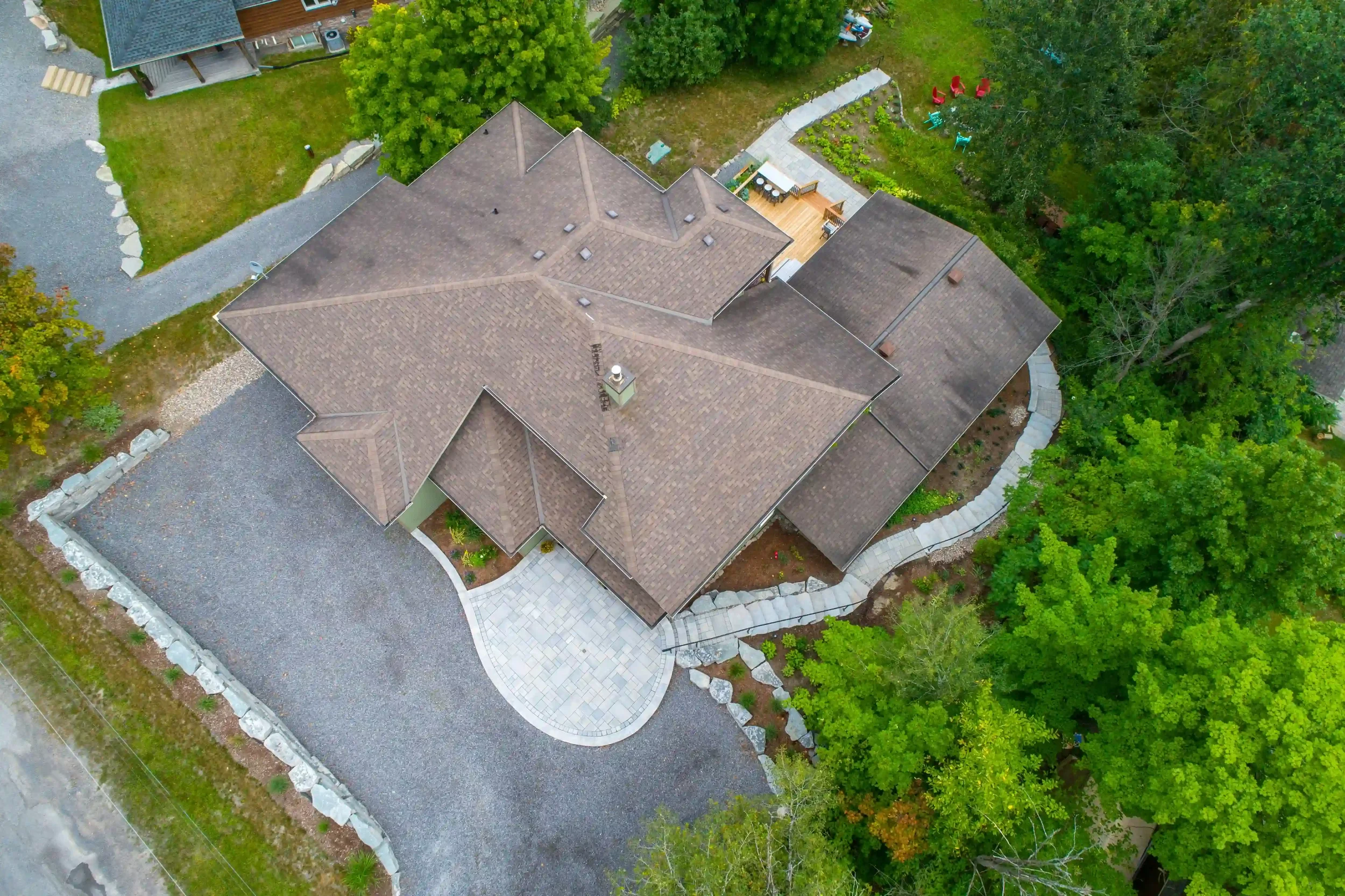 An aerial view of a house with a complex roof design, surrounded by trees and a landscaped yard with paved walkways and a gravel driveway.