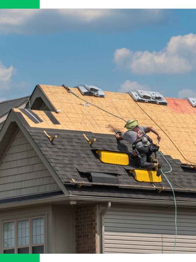 A contractor working on the roof of a house, installing new shingles while hanging from the roof with safety harnesses and ropes during daytime.