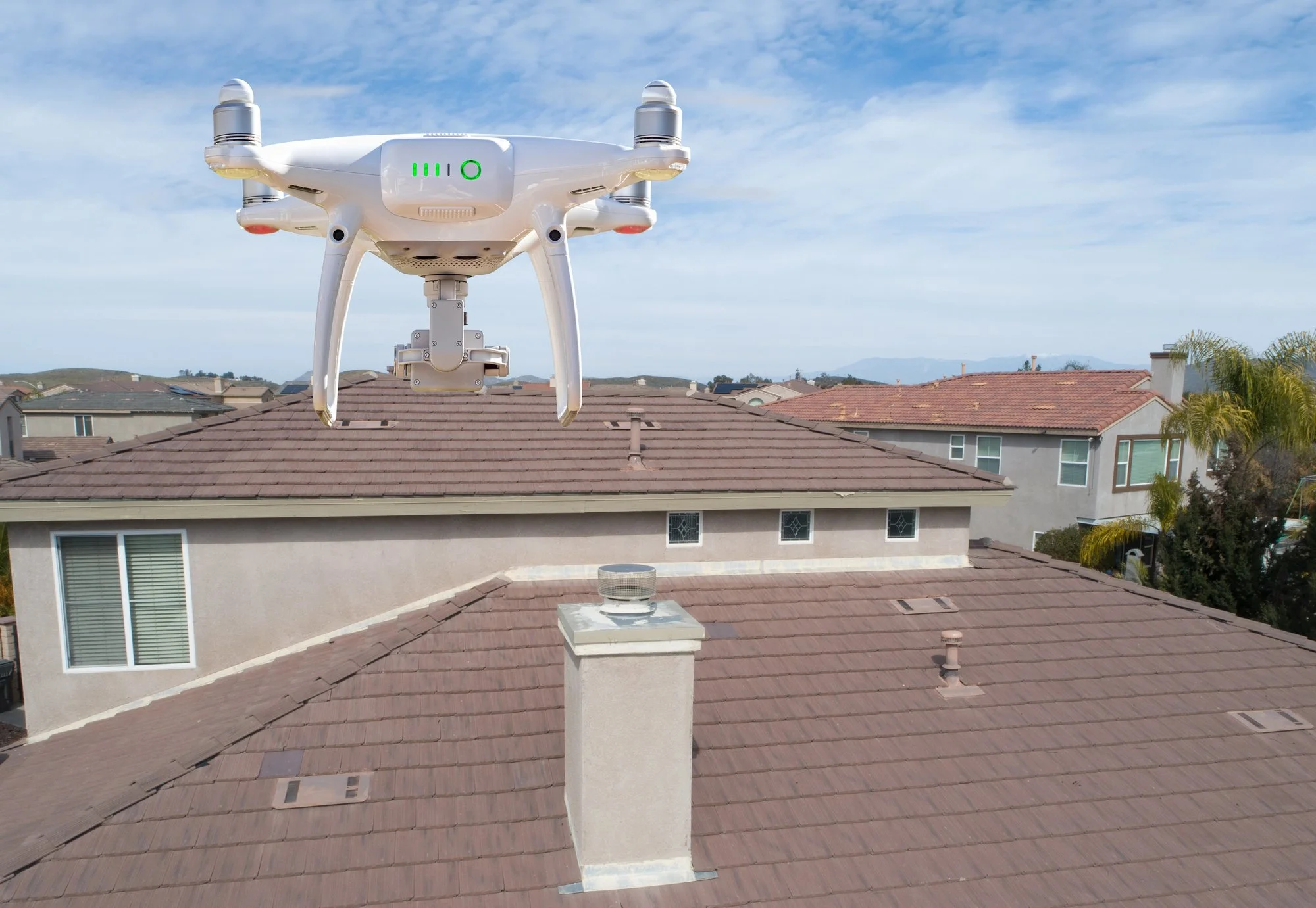 A drone hovering above rooftop residential houses under partly cloudy blue sky.