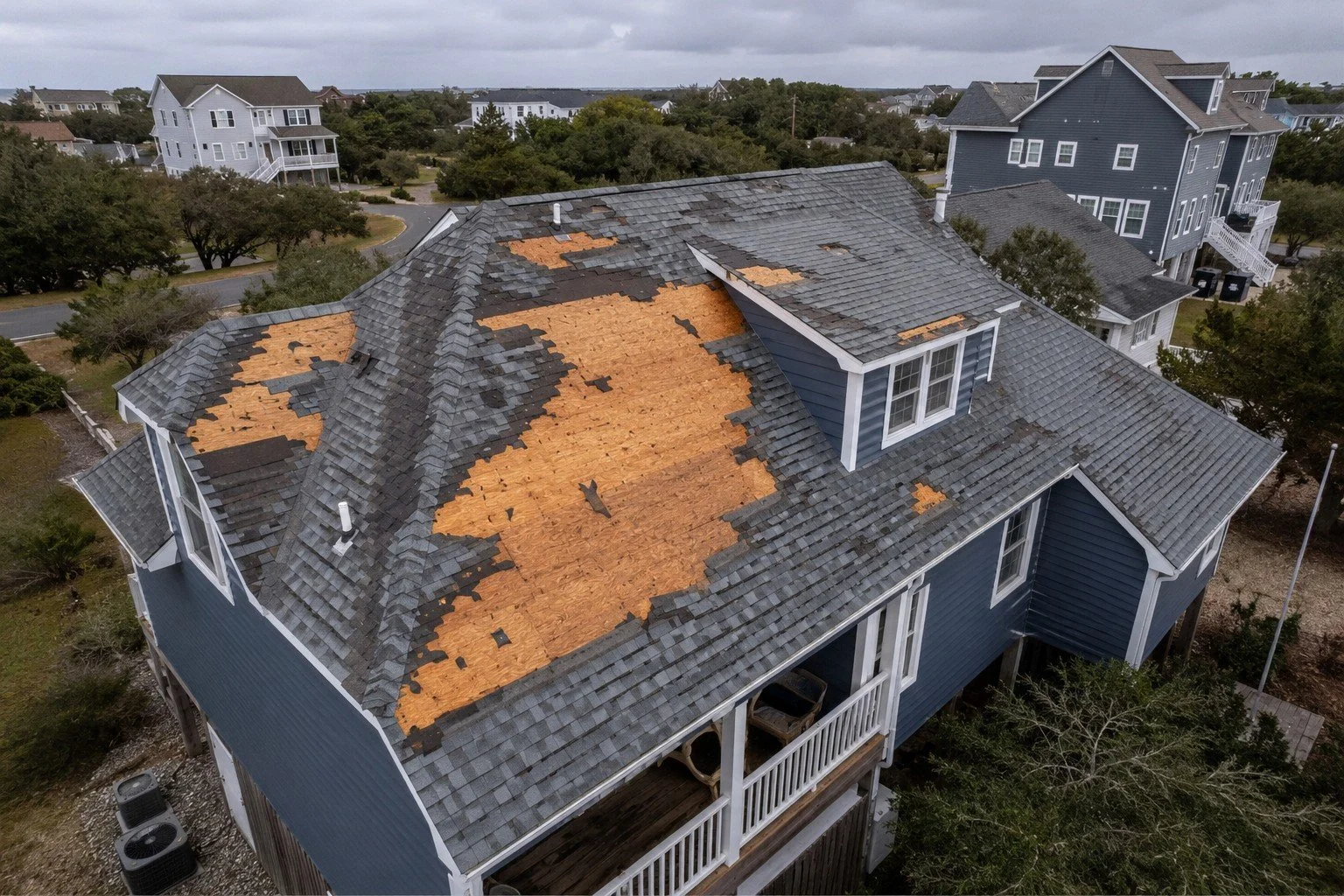 Aerial view of a house with a blue exterior and multiple gabled roofs, showing extensive shingle damage with sections of missing and damaged roofing materials.