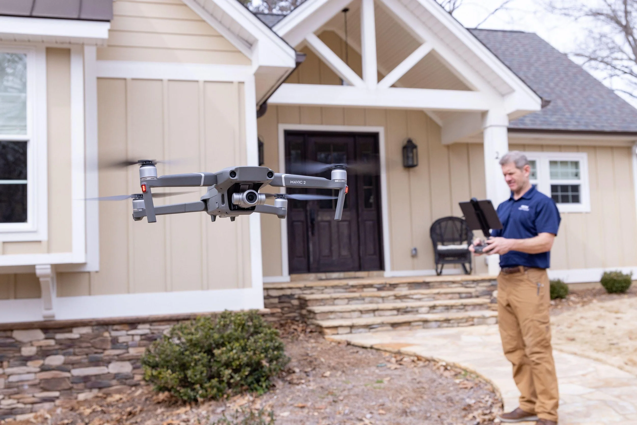 A man is operating a drone in front of a suburban house with a beige exterior and stone steps. The drone is flying in the air near him.