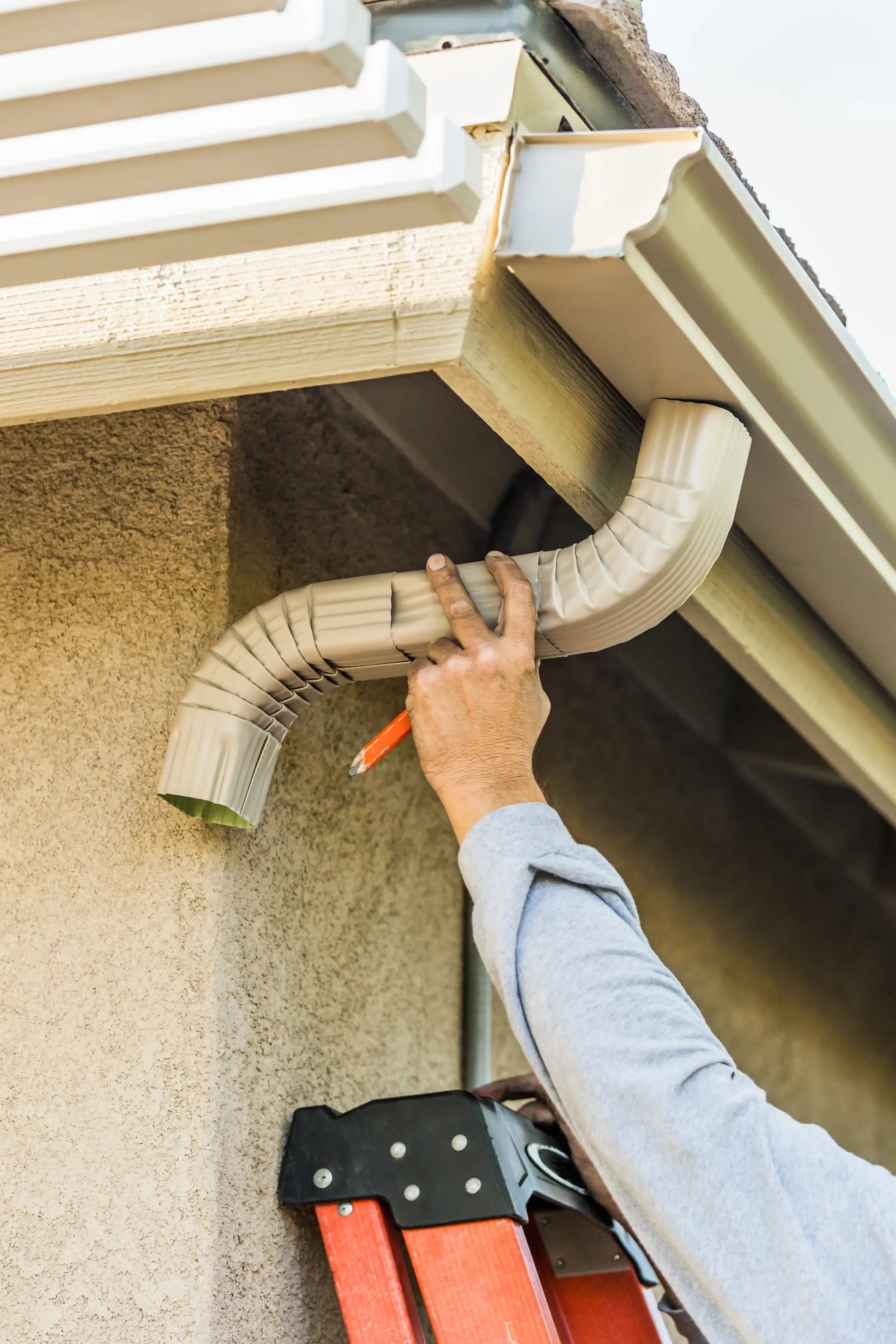 A person installing or repairing a vent or duct at the corner of a building's exterior wall, using a step ladder and holding a tool.