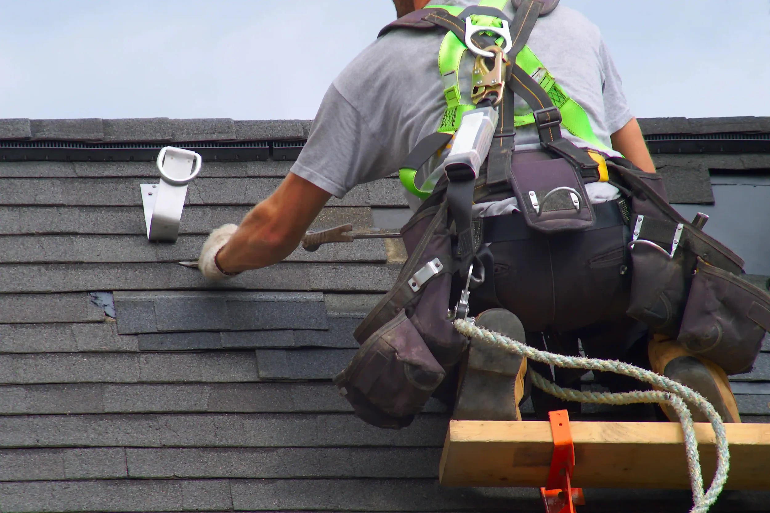 A worker installing or repairing a shingled roof, wearing safety harness and yellow protective boots, using a hammer and working with shingles.