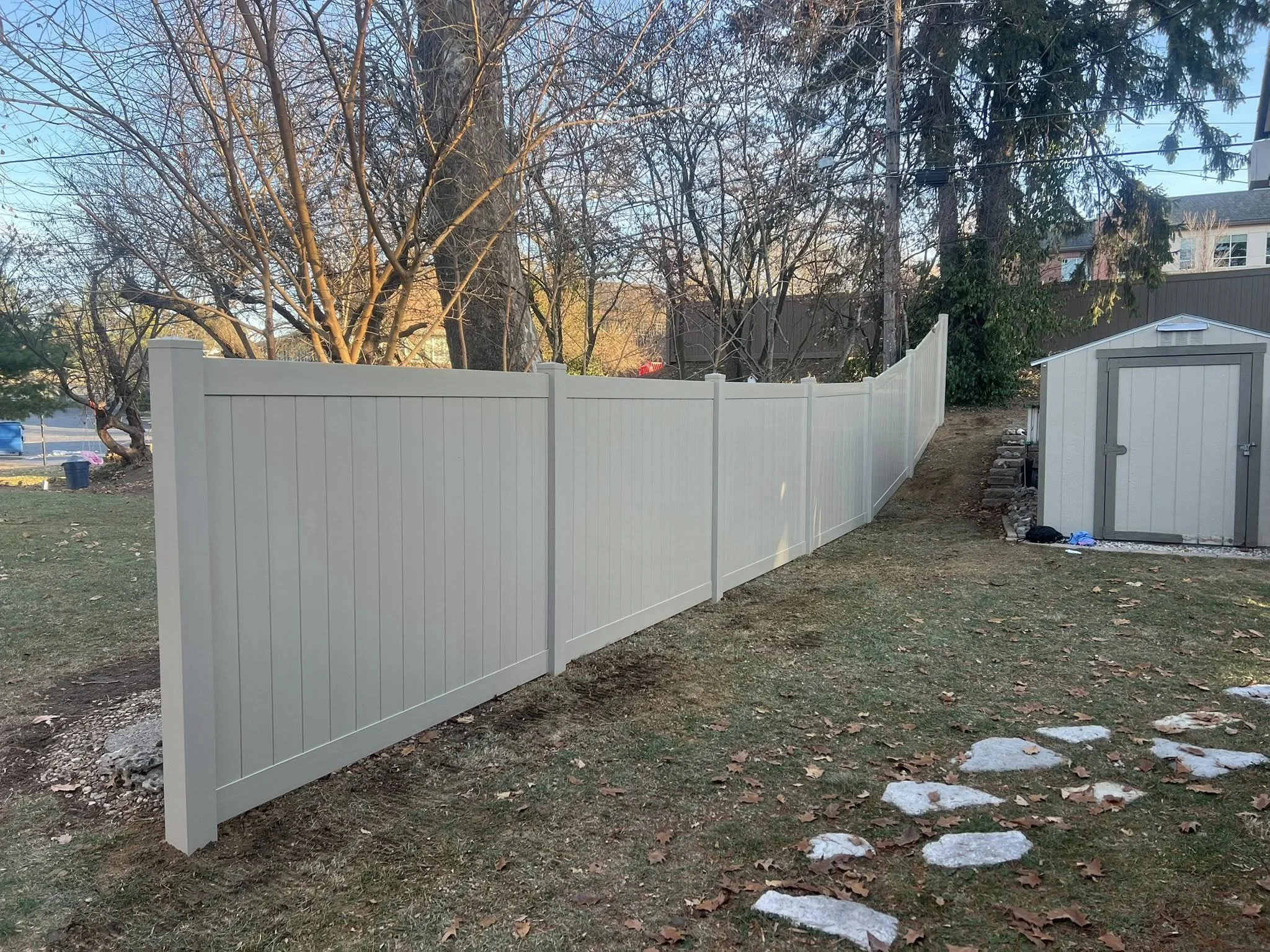 A beige vinyl privacy fence with vertical panels running alongside a yard. There are trees, a small beige shed, and a slope covered with dirt and rocks. The setting appears to be in a suburban backyard during late fall or winter.