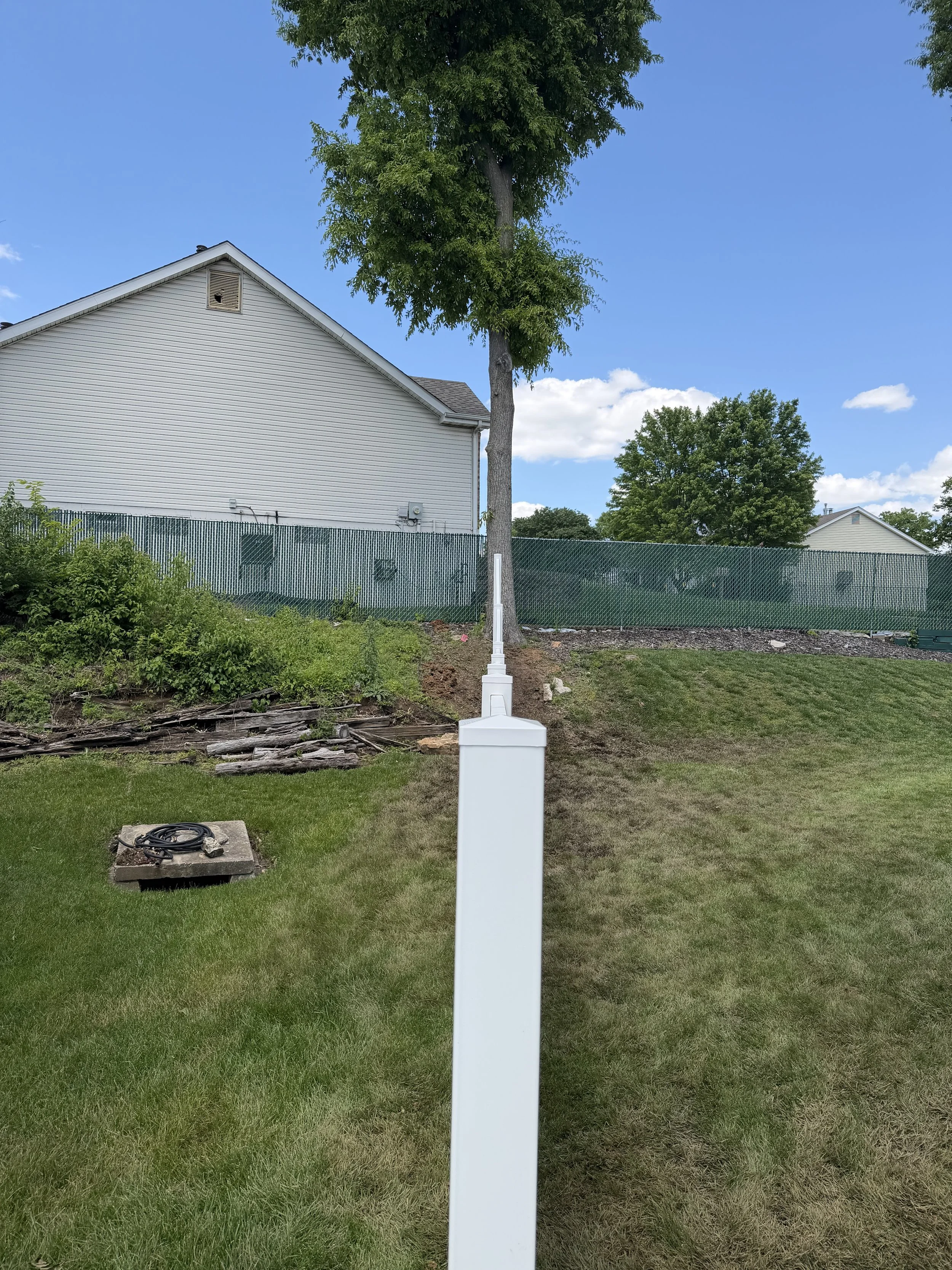 A white vertical post installed in a grassy yard. Behind it, a tall leafy tree, a white house with beige siding, a chain-link fence, and additional trees are visible under a blue sky with scattered clouds.