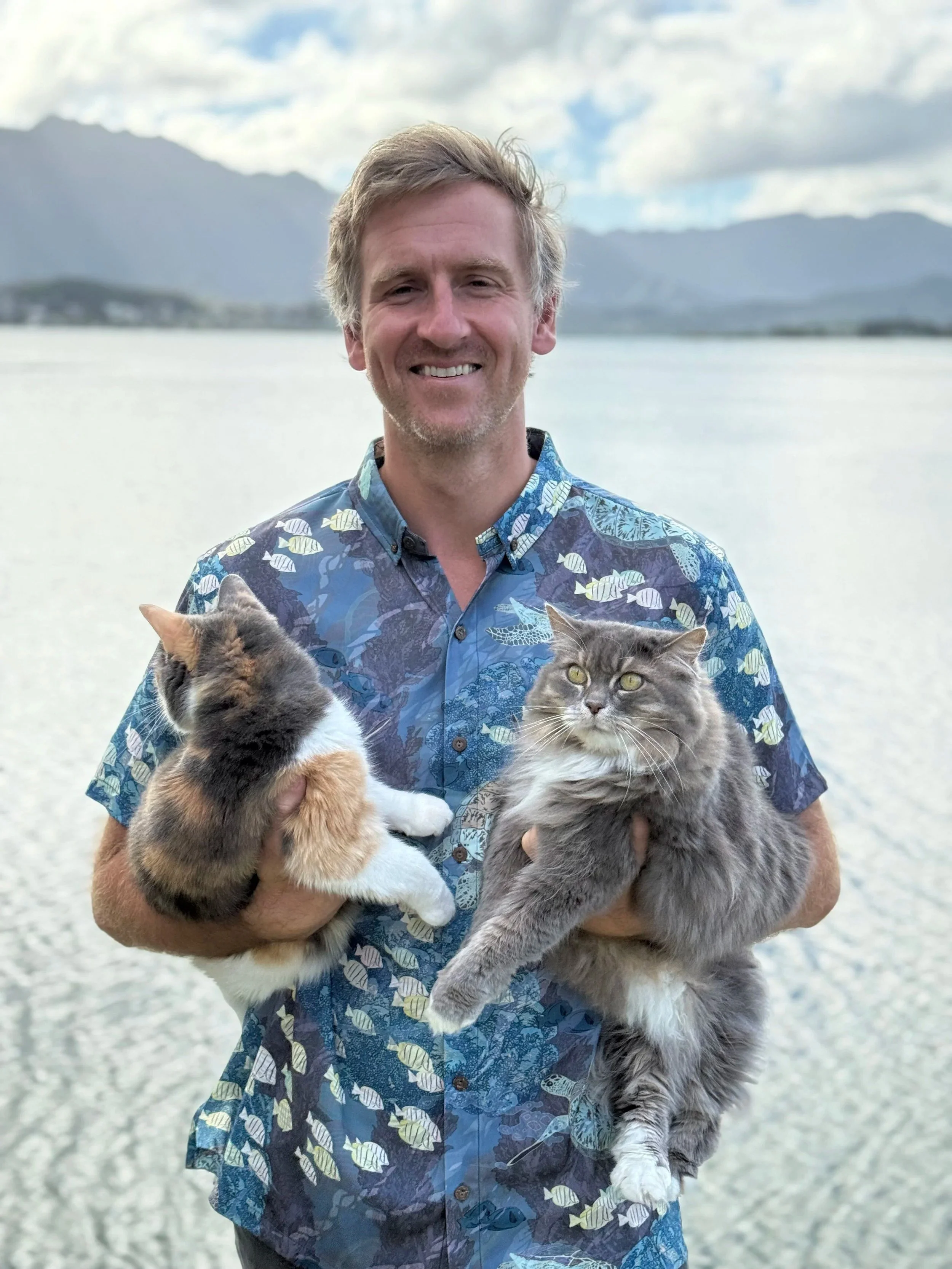 A smiling man holding two cats, one calico and one gray, near a body of water with mountains and a cloudy sky in the background.