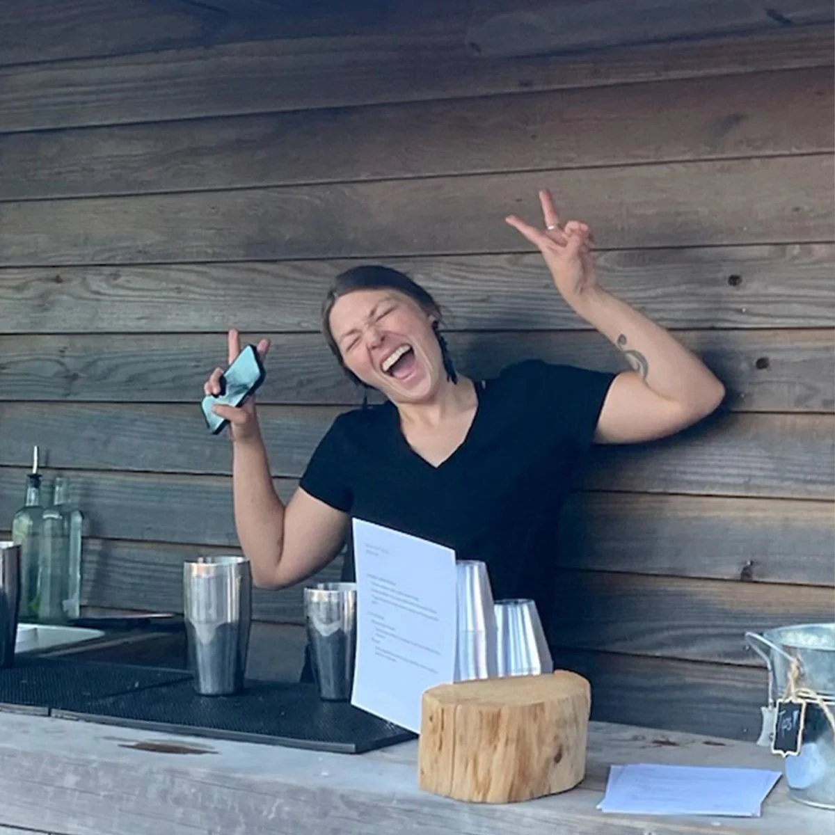 A woman with short dark hair, wearing a black t-shirt, joyfully singing or shouting with eyes closed. She is holding a smartphone in her right hand and making a peace sign with her left hand while sitting at a table with glasses and a wooden block against a wooden wall background.