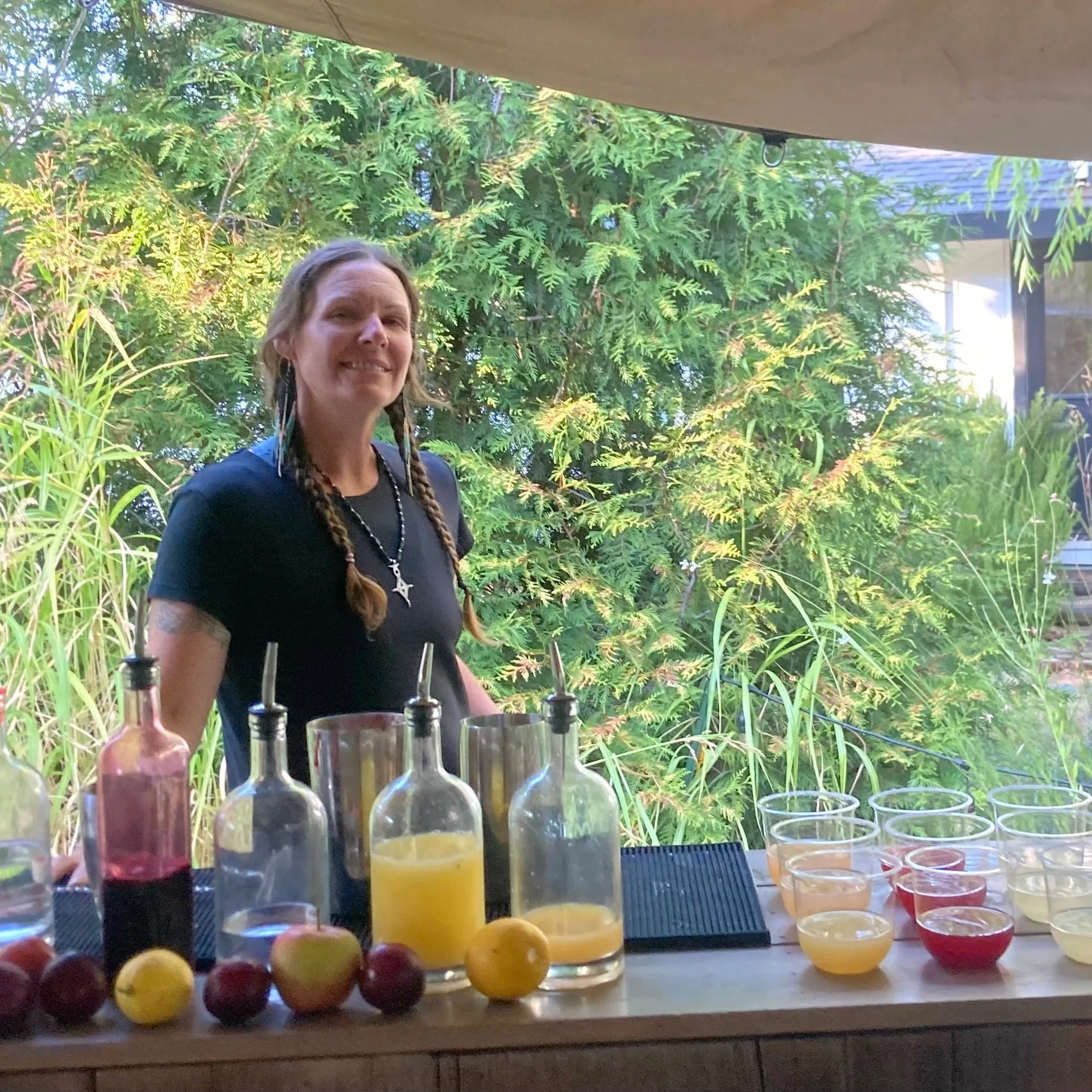 A woman with braids and necklaces standing behind a table with bottles and glasses filled with different colored beverages, outdoors with green foliage in the background.