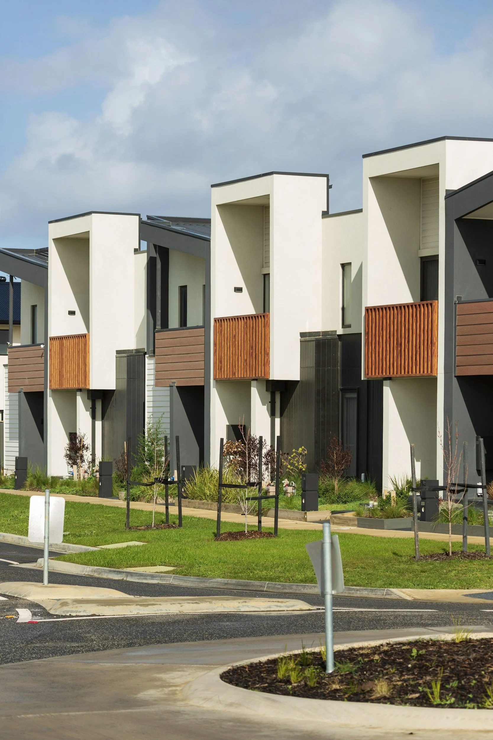 Modern white residential buildings with wooden accents, small landscaped gardens, and a curved sidewalk in the foreground, under a partly cloudy sky.