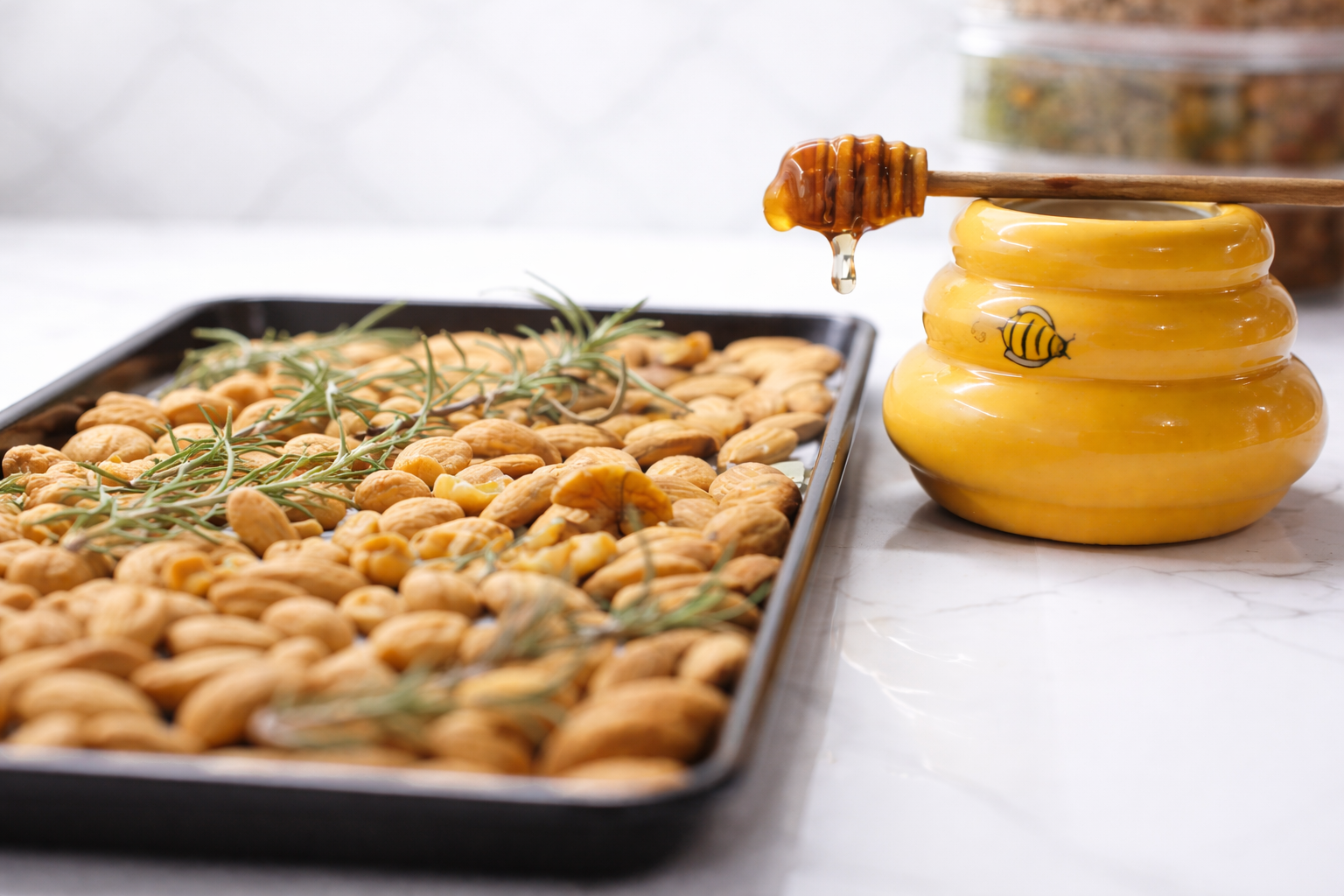 Tray of roasted nuts garnished with rosemary next to a yellow honey jar with honey on a honey dipper