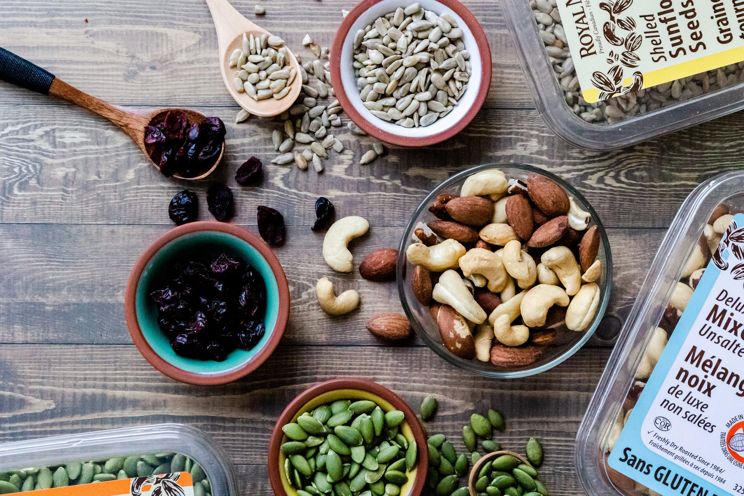 Various containers and bowls filled with seeds, nuts, dried cranberries, and green beans on a wooden surface.