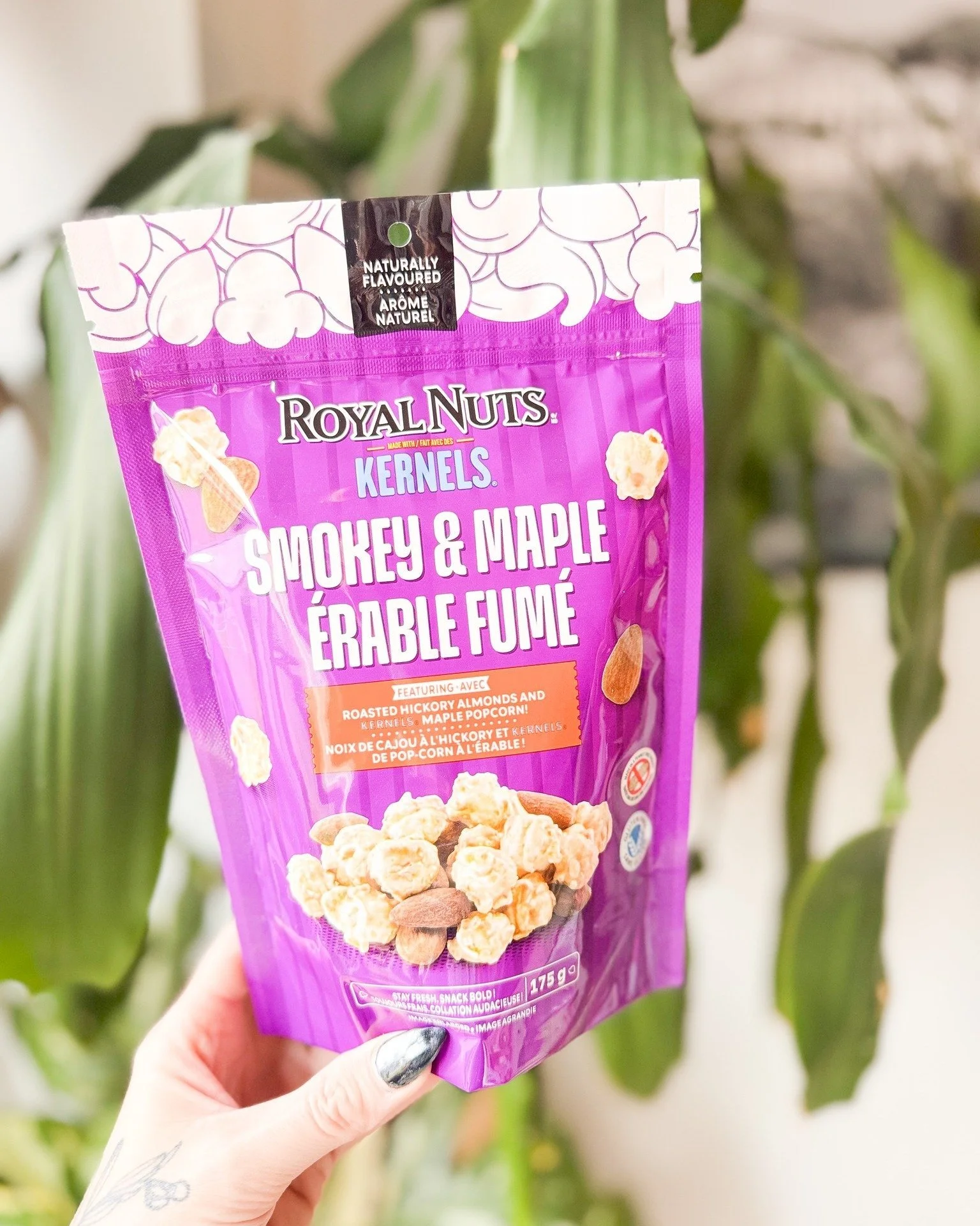 Purple packaging of Royal Nuts Smoky & Maple Kernels popcorn held by a hand with painted nails, in front of green leaves.