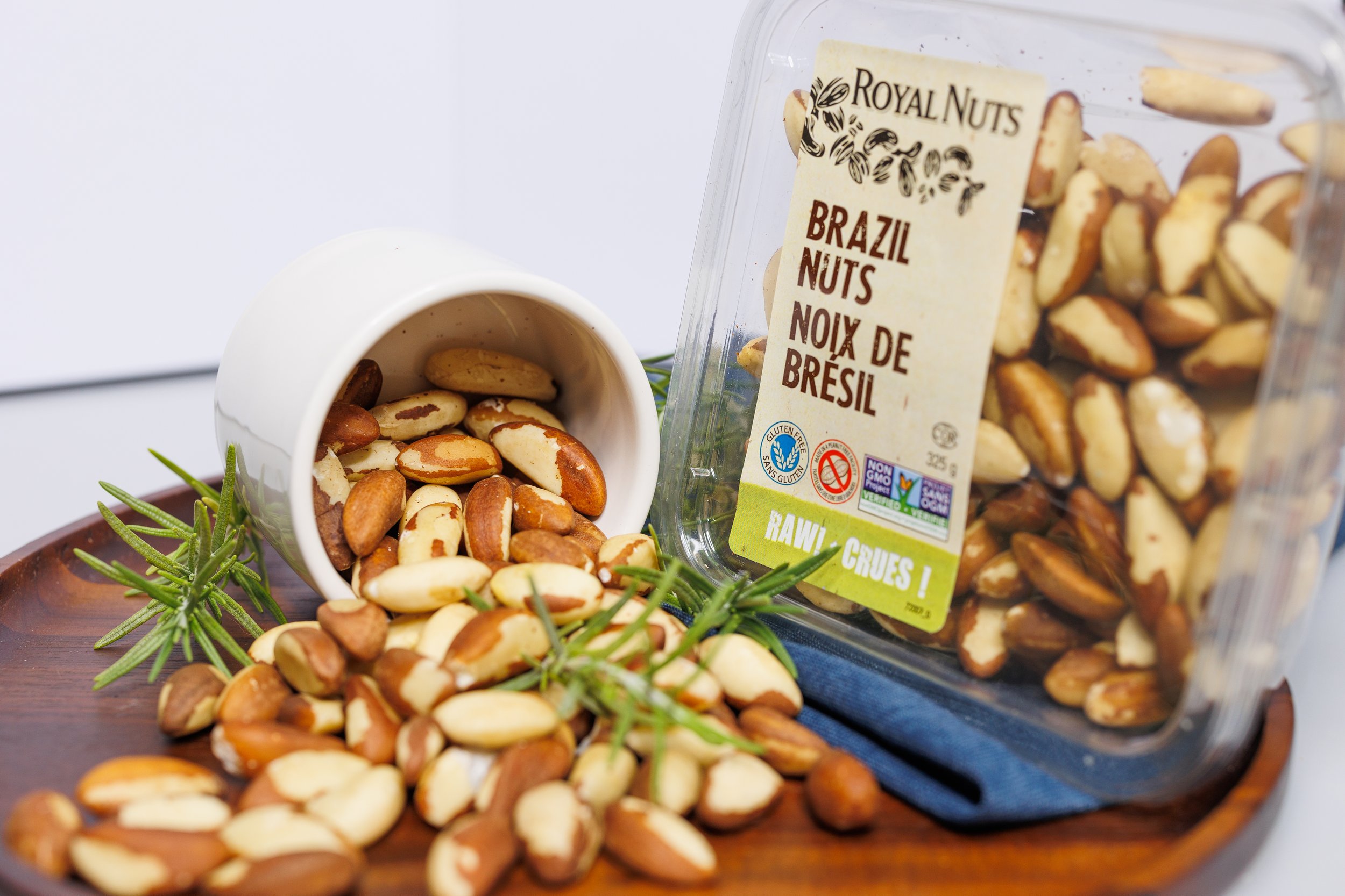 A clear plastic container of Brazil nuts with a label indicating they are raw and from Brazil, partially open with some nuts visible inside. A white cup filled with Brazil nuts is overturned next to the container. The scene includes a small sprig of green herbs or leaves on a wooden surface.