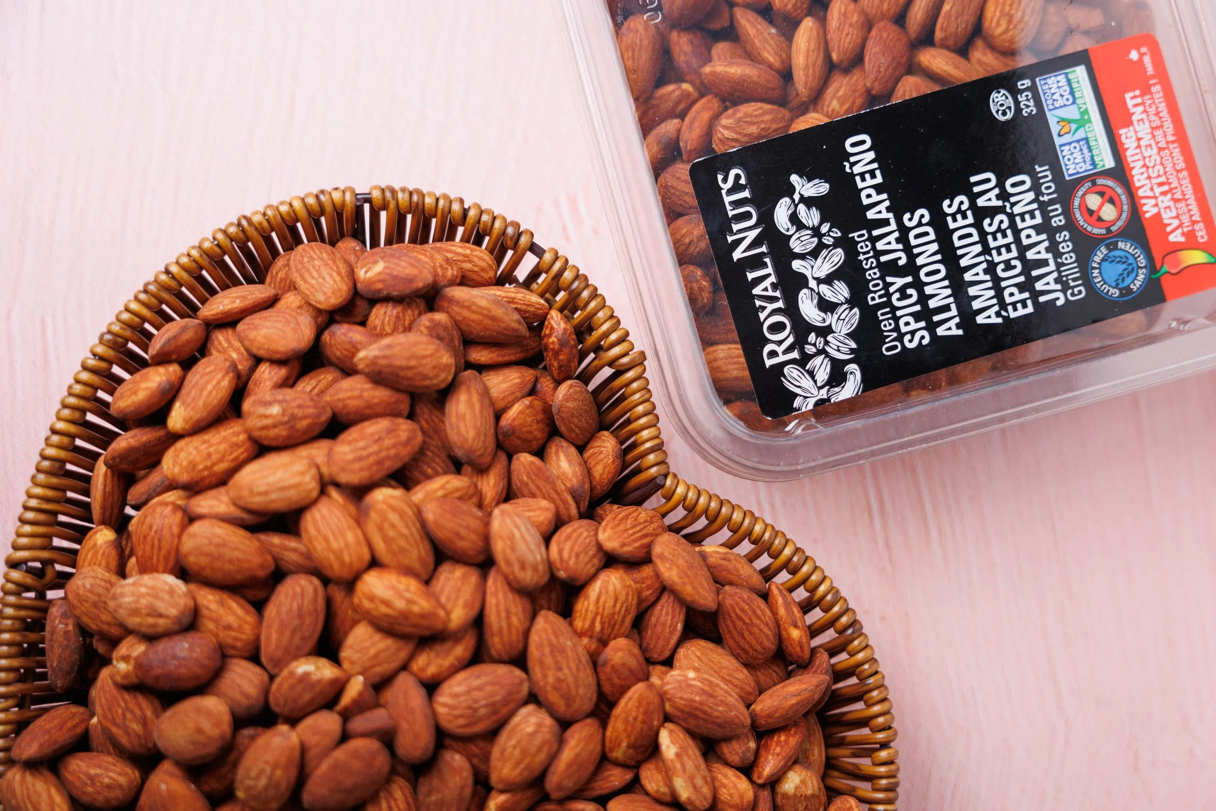 A clear plastic container of roasted almonds with a black and red label, placed next to a woven basket filled with almonds on a pink wooden surface.