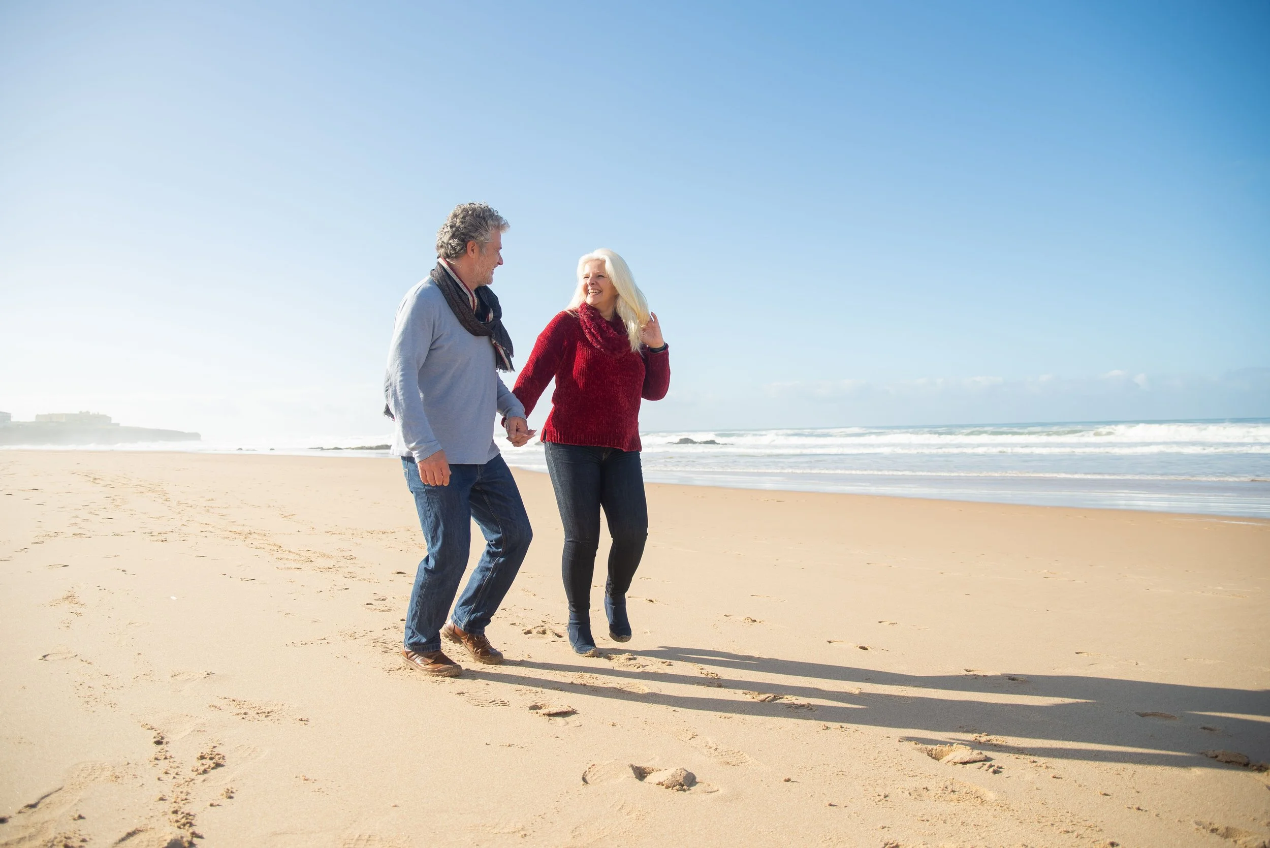 A man and a woman walking hand in hand along the sandy beach with the ocean and blue sky in the background.