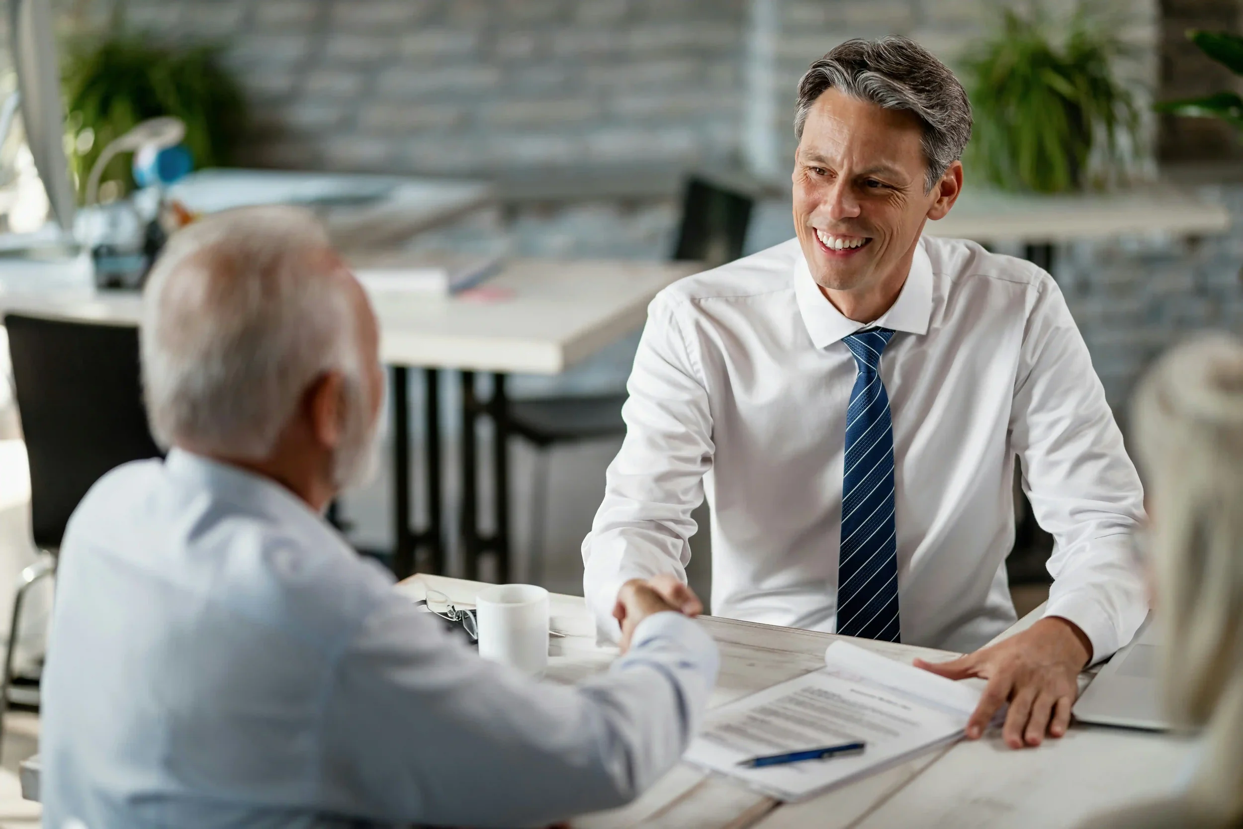 Two men, one younger with dark hair and one older with white hair, are sitting at a table in an office or meeting room. They are engaged in a friendly conversation, smiling and shaking hands. The younger man is wearing a white shirt and a blue tie, while the older man is also dressed in a white shirt. The background shows some office equipment and plants, indicating a professional setting.
