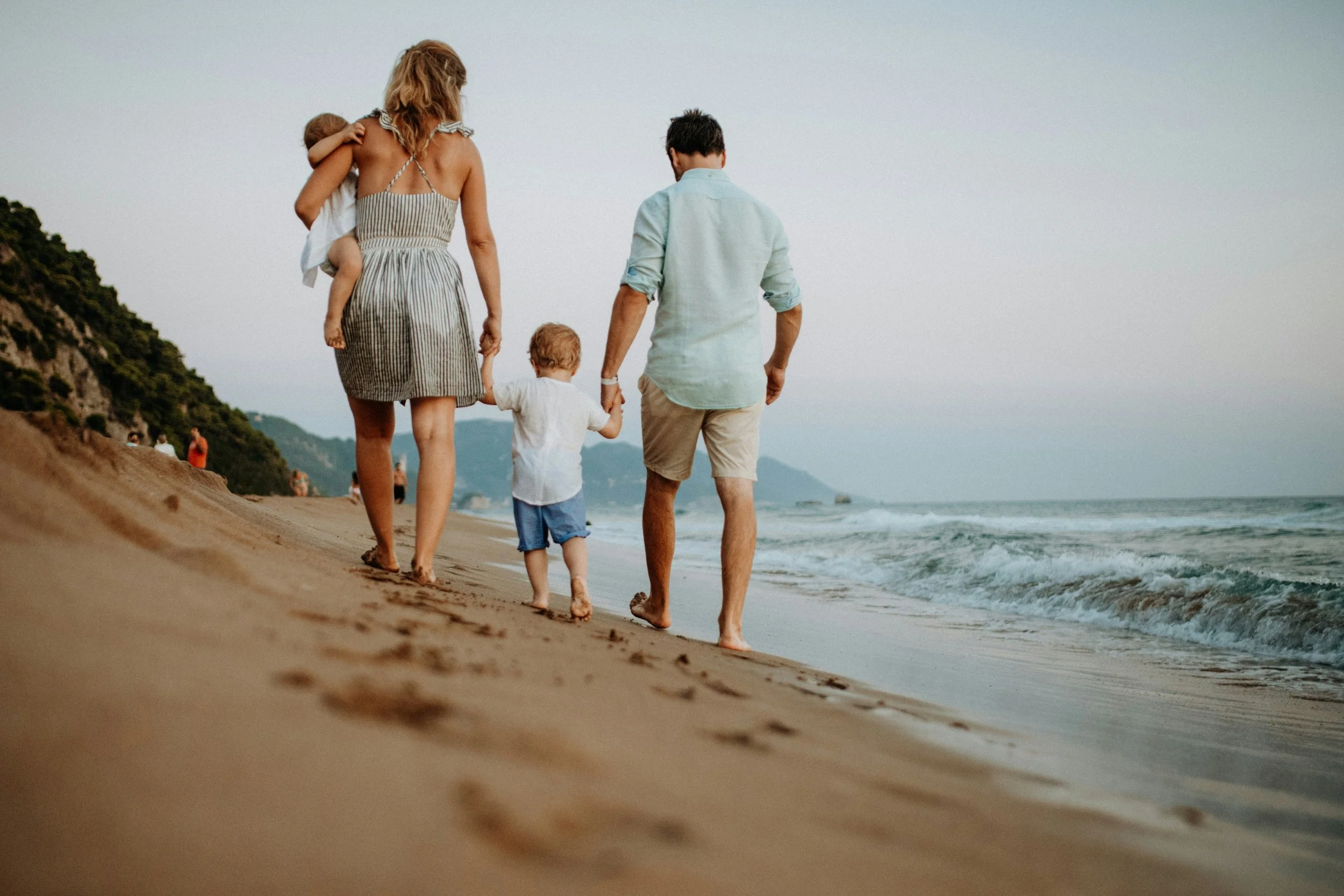 A family walking on the beach at sunset, with one woman carrying a child and holding hands with two young boys, all facing the ocean.