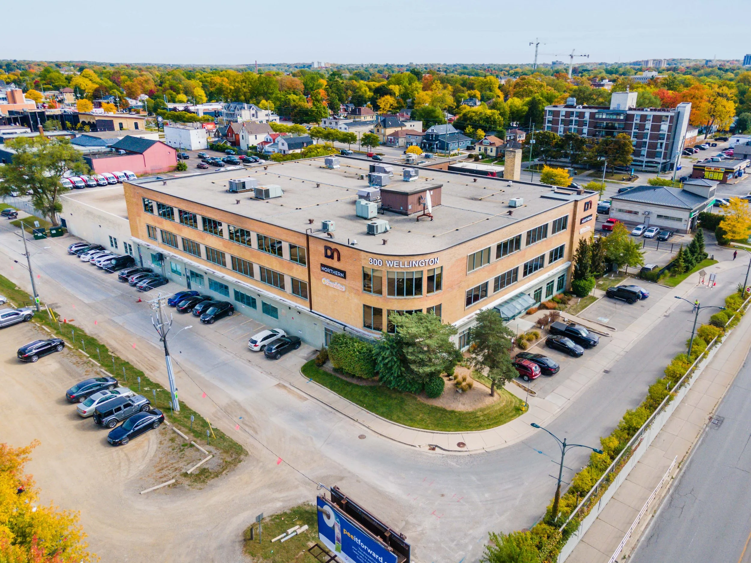 Aerial view of a multi-story commercial building at 300 Wellington with parking lots, surrounding greenery, and nearby residential and commercial buildings.