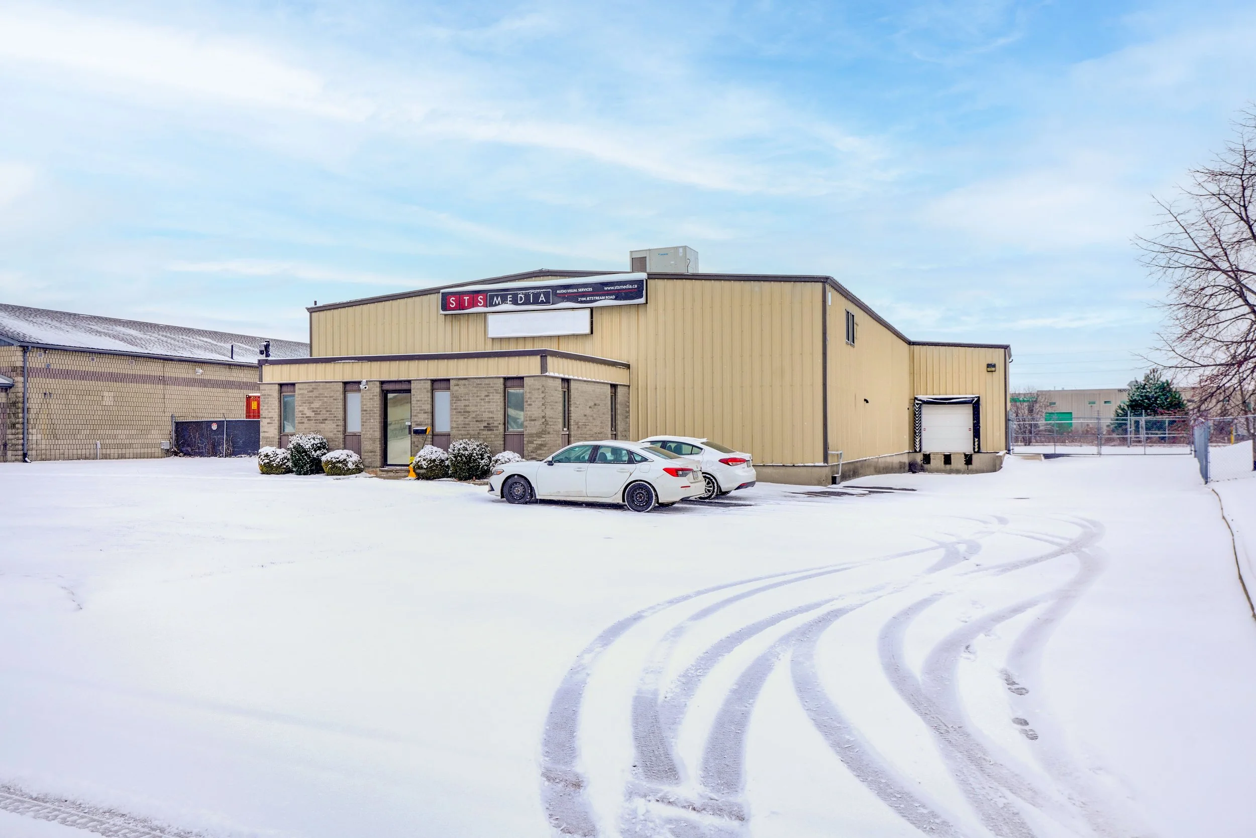 Snow-covered parking lot in front of a commercial building with parked cars, a sign that says 'STS Media', and a clear blue sky.