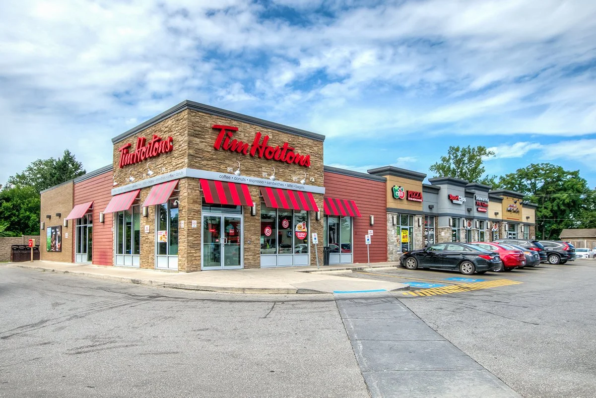 A city street scene with a small two-story brick building in the foreground, surrounded by high-rise residential buildings and construction cranes in the background. Cars are parked along the street, and utility poles with wires are visible.
