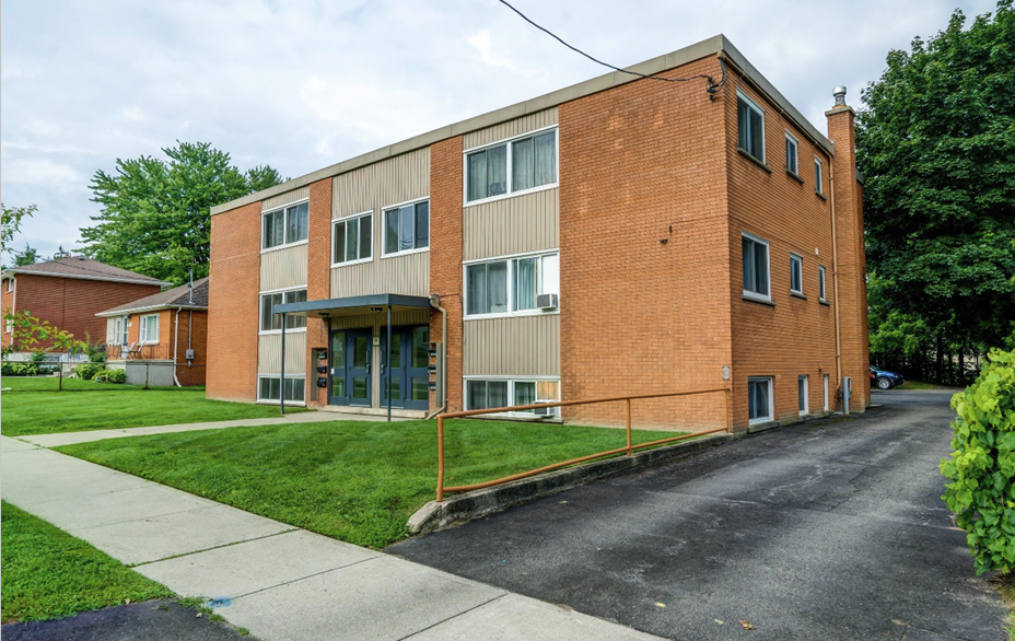 Three-story brick apartment building with beige siding accents, blue doors, and multiple windows, surrounded by a green lawn and sidewalk.