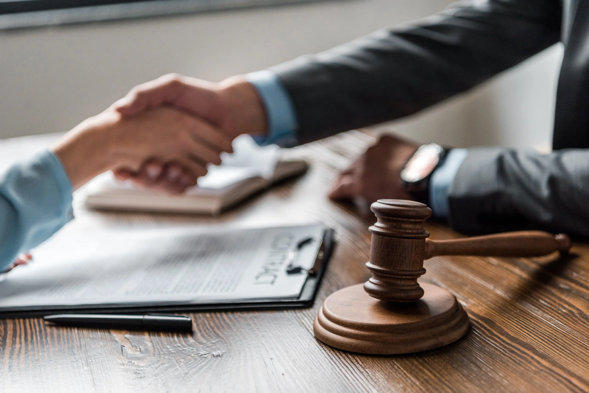 Two people shaking hands over a wooden table, with a chess pawn in the foreground and legal documents and a pen on the table.