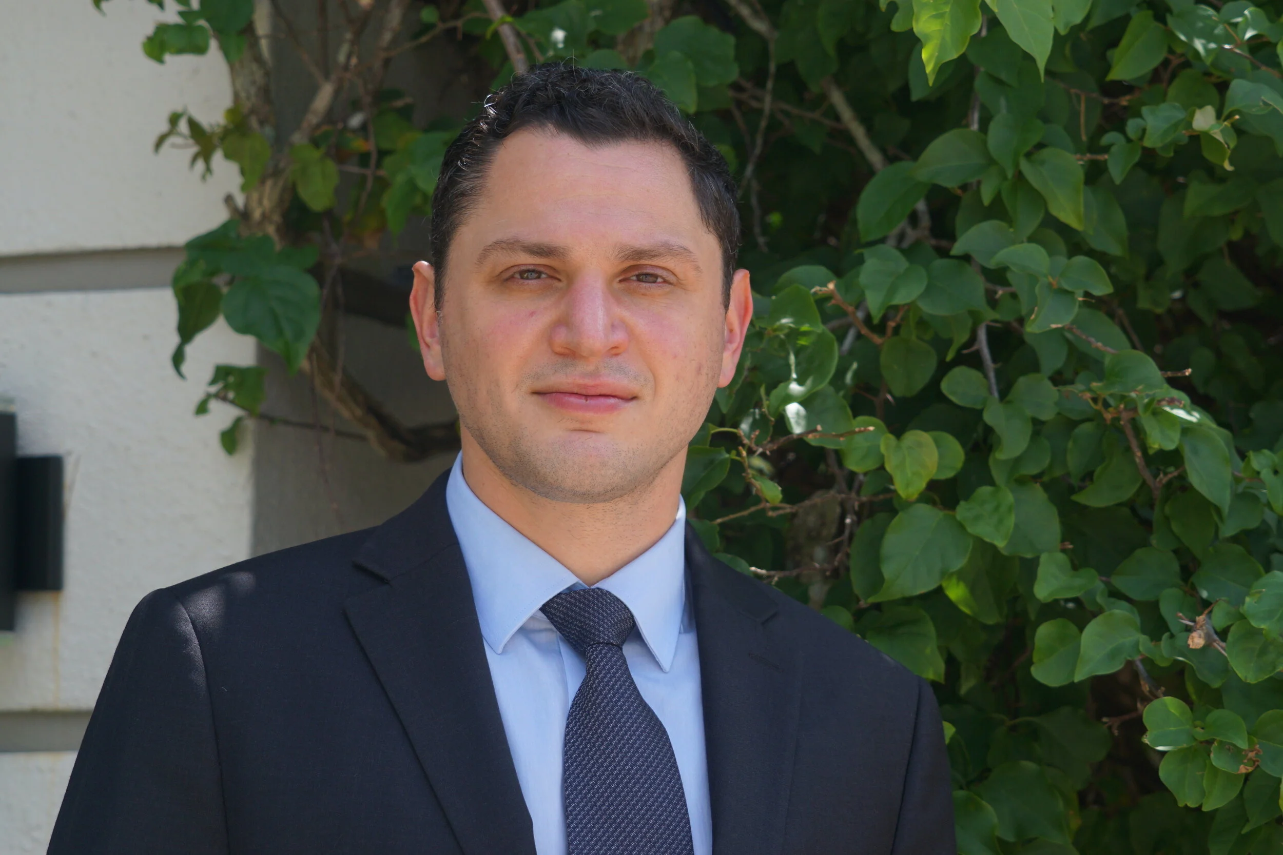 Man in formal suit and tie standing outdoors with green leafy background.