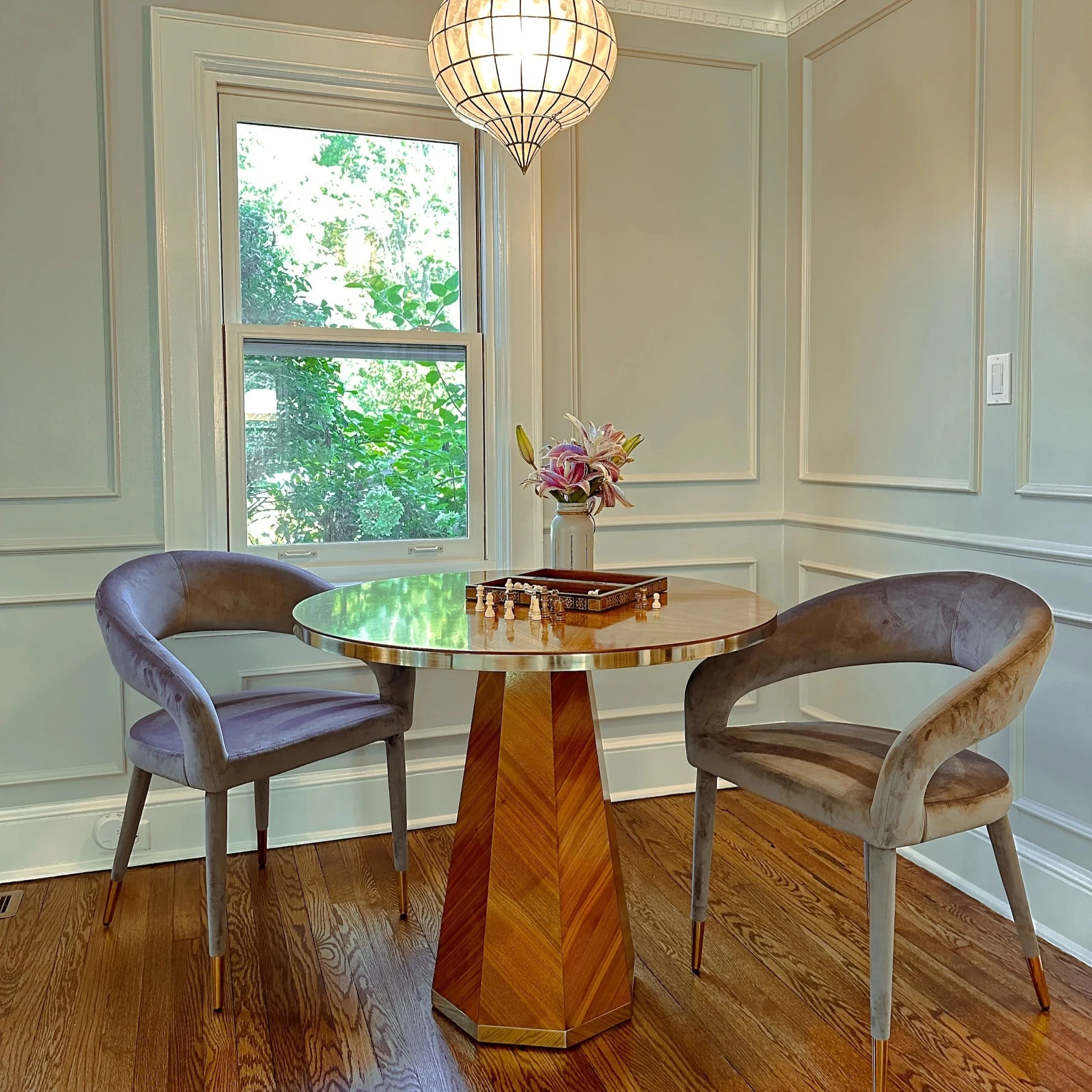 A cozy dining nook with a round wooden table, two velvet chairs, a window with lush greenery outside, a vase with pink lilies, and a pendant light overhead in an elegant corner of a room.
