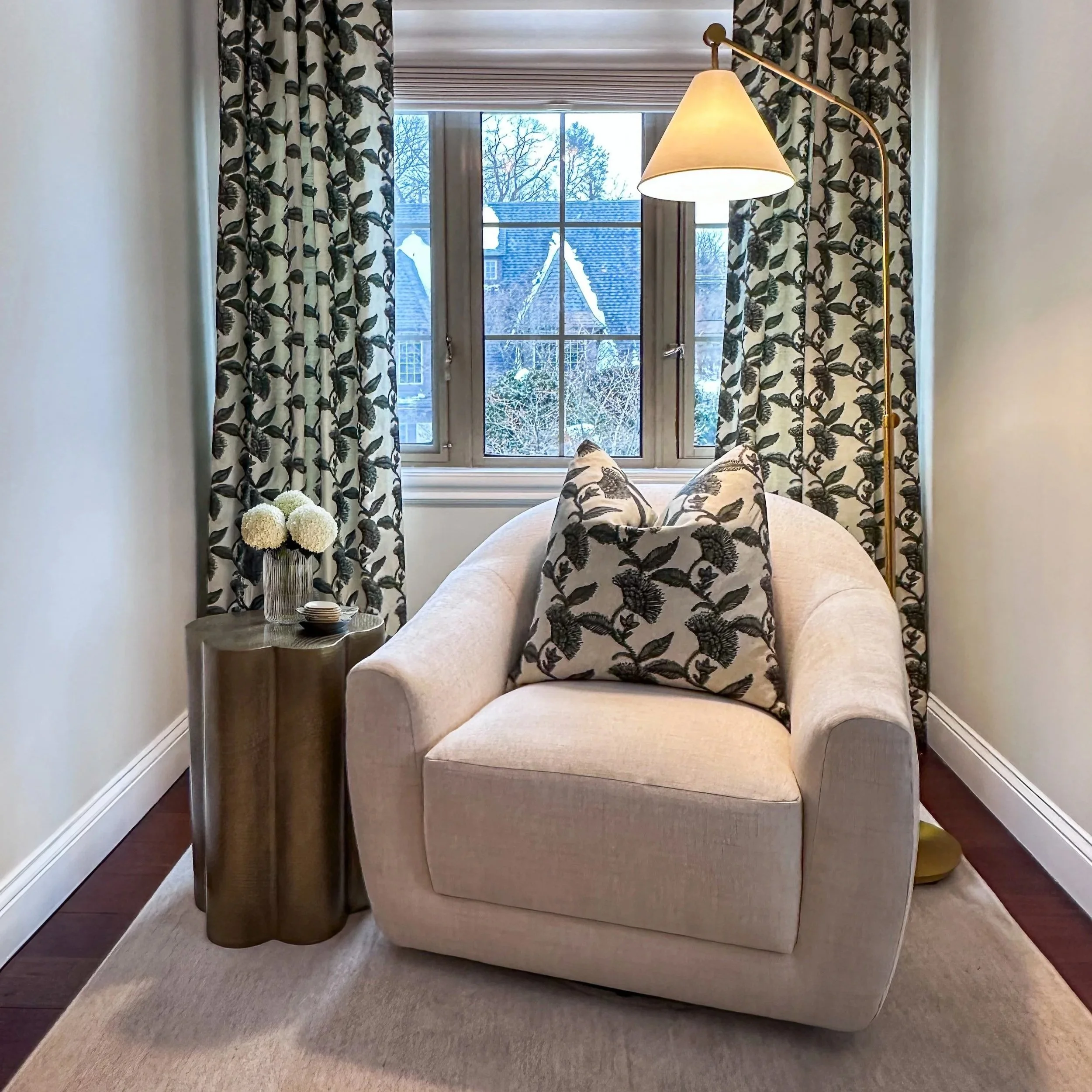A cozy sitting area with a beige armchair, two decorative pillows with a black and white leafy pattern, a small side table with a flower vase, and a window with matching patterned curtains. A floor lamp with a white lampshade is placed beside the chair, and outside the window, snow-covered rooftops are visible.