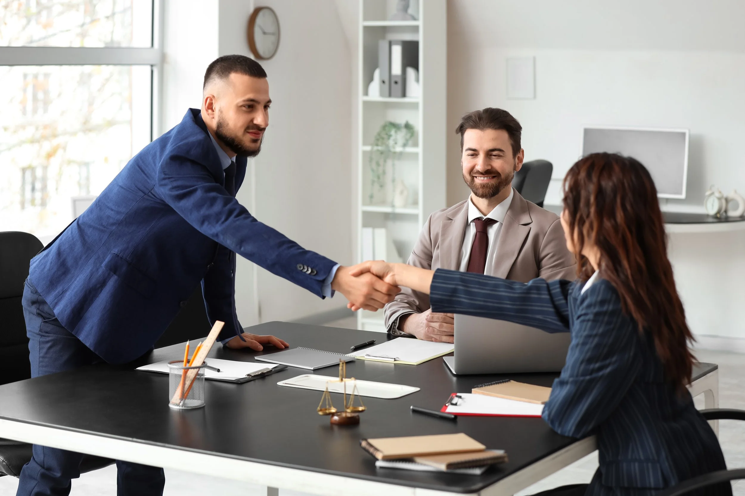 Three business professionals in an office, two men and one woman, engaged in a handshake over a meeting table, with notebooks, pens, and a scale model on the table.